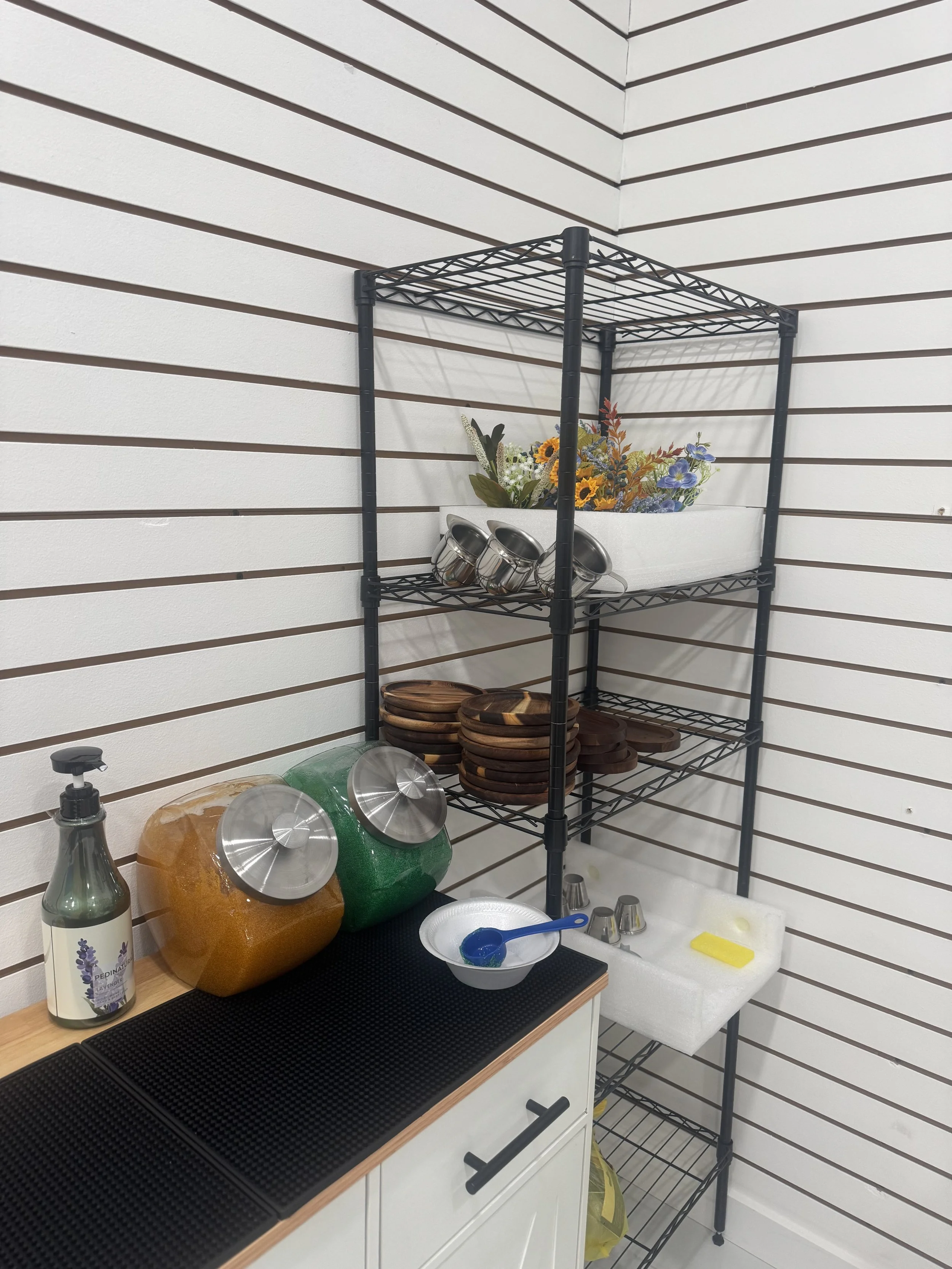 Kitchen corner with a black wire shelving unit, a white flower box with flowers, several small metal cups, a countertop with a bottle of lavender hand soap, large decorative jars, and a white dish with a blue spoon, yellow sponge, and cleaning suppli