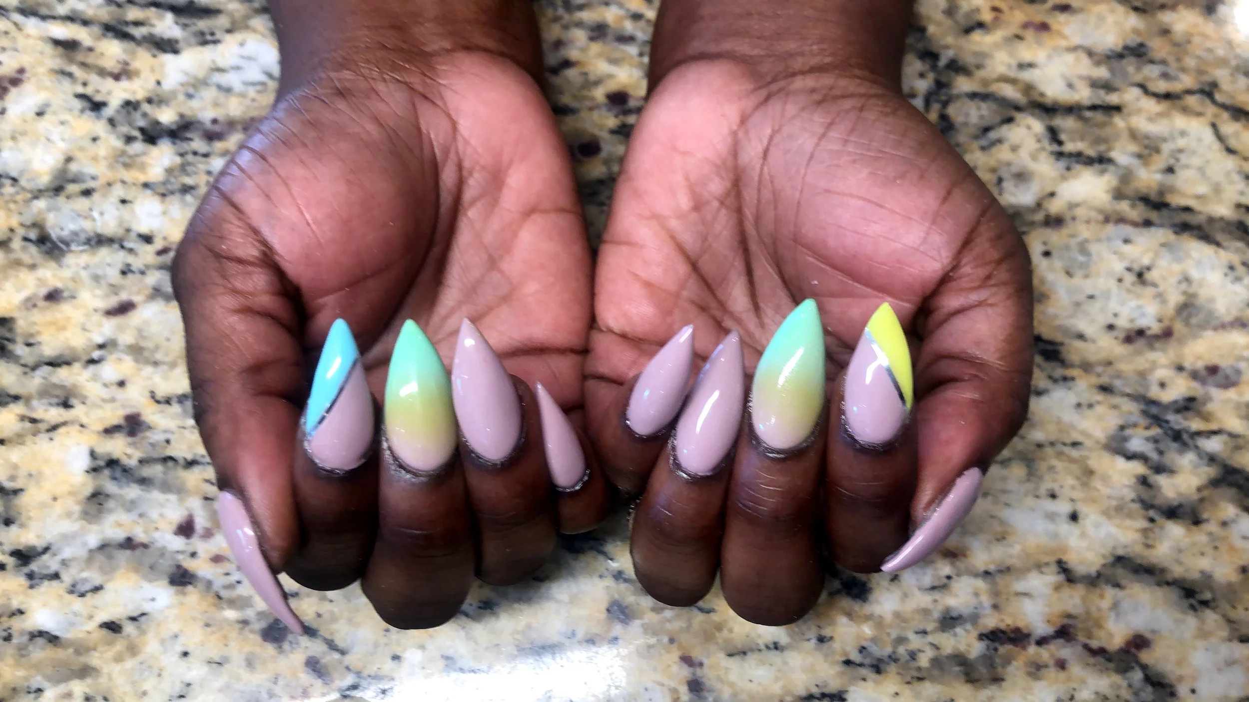A pair of hands with long, pointed nails painted in pastel colors with black accents, resting on a speckled granite surface.