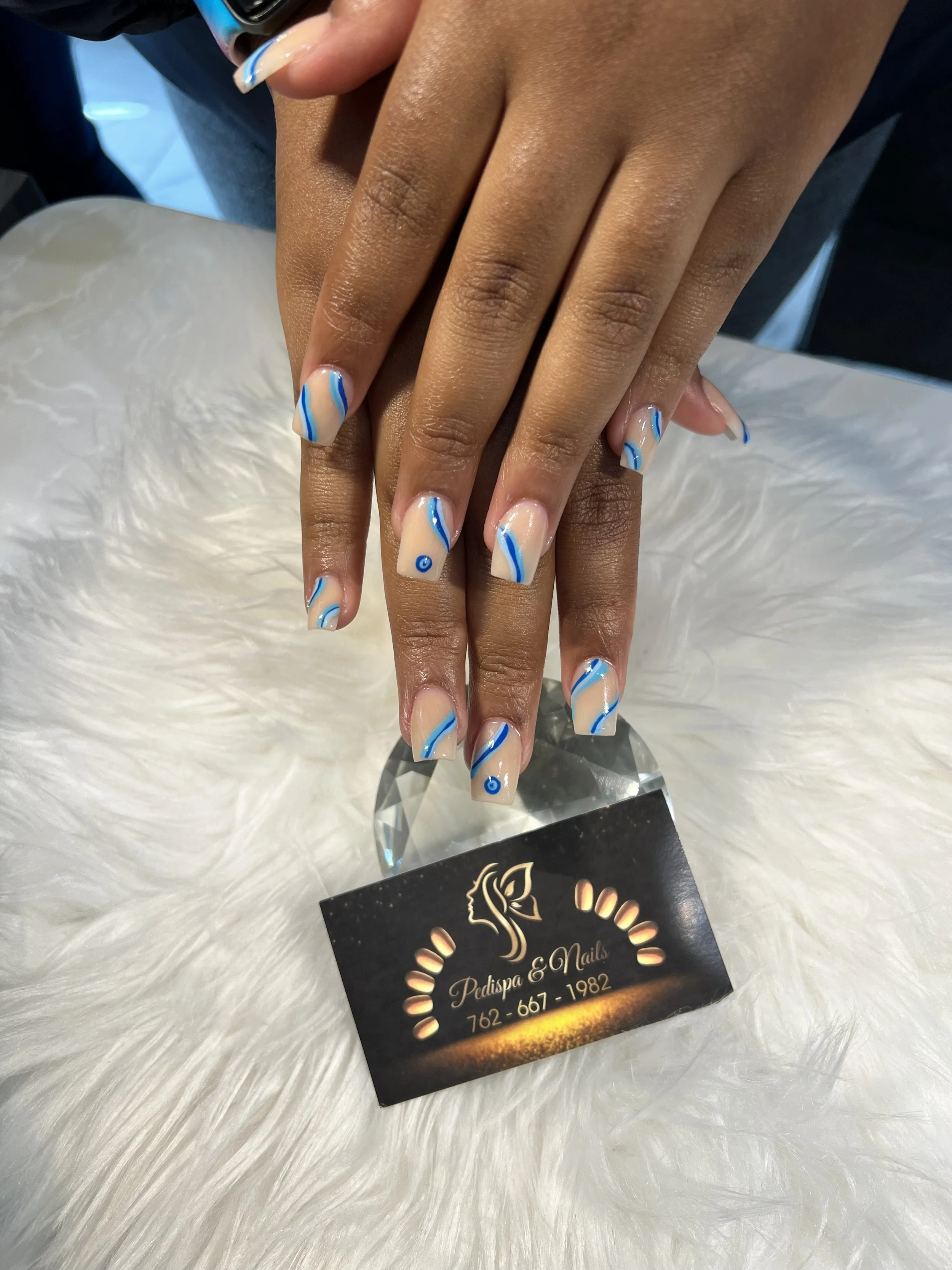 Hands with blue and white nail art resting on a white furry surface near a black card with gold writing.