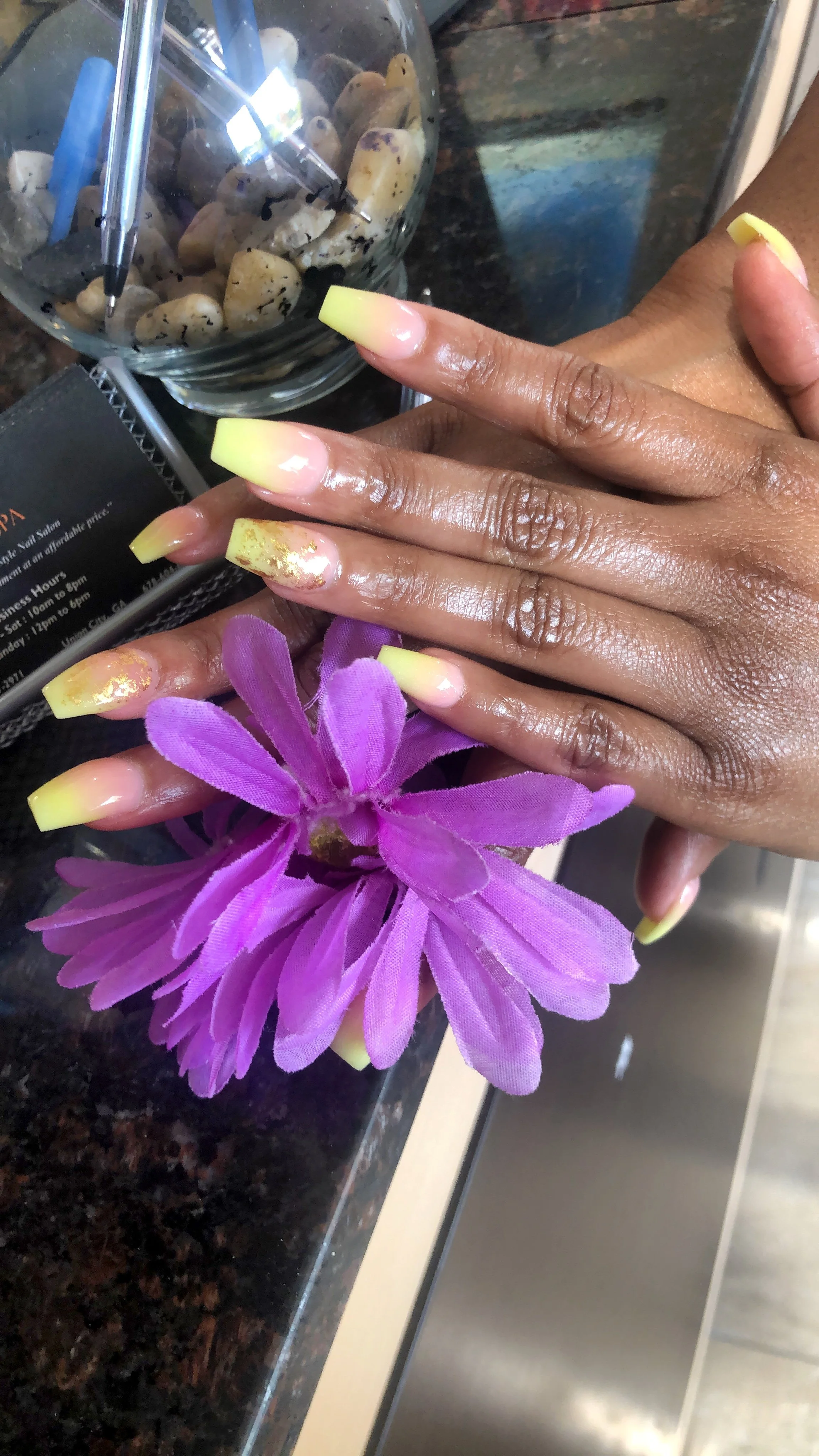 A person's hand with long yellow and pink acrylic nails resting on a purple artificial flower on a dark granite counter. In the background, there is a glass container filled with rocks and two blue pens.