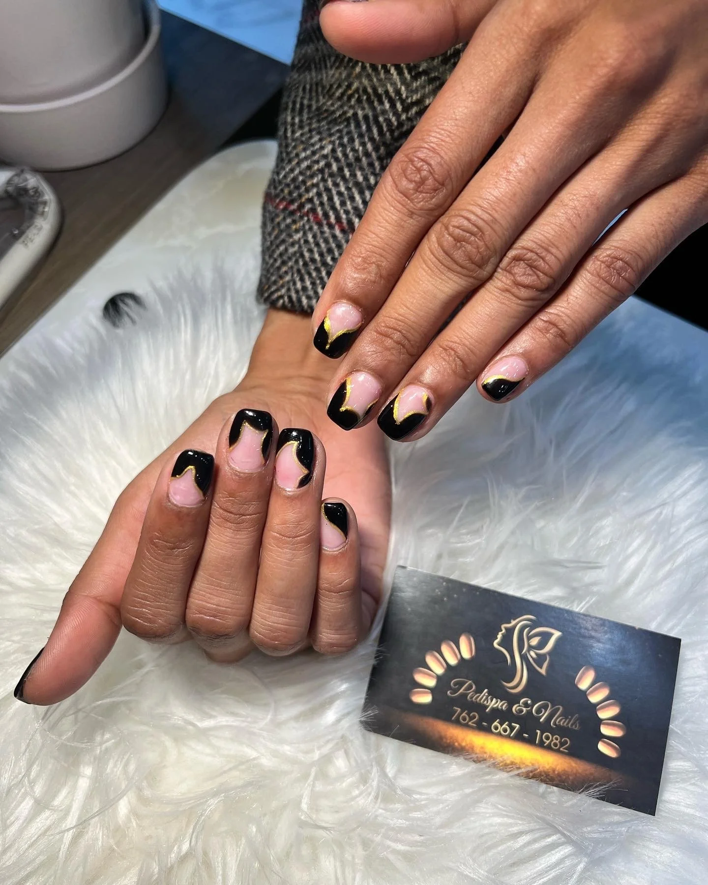 Close-up of a person's hand with a fresh black and gold manicure, resting on a white furry surface. Another hand is gently touching the first hand. In the background, part of a business card for Pedicure & Nails is visible, displaying a logo, phone n