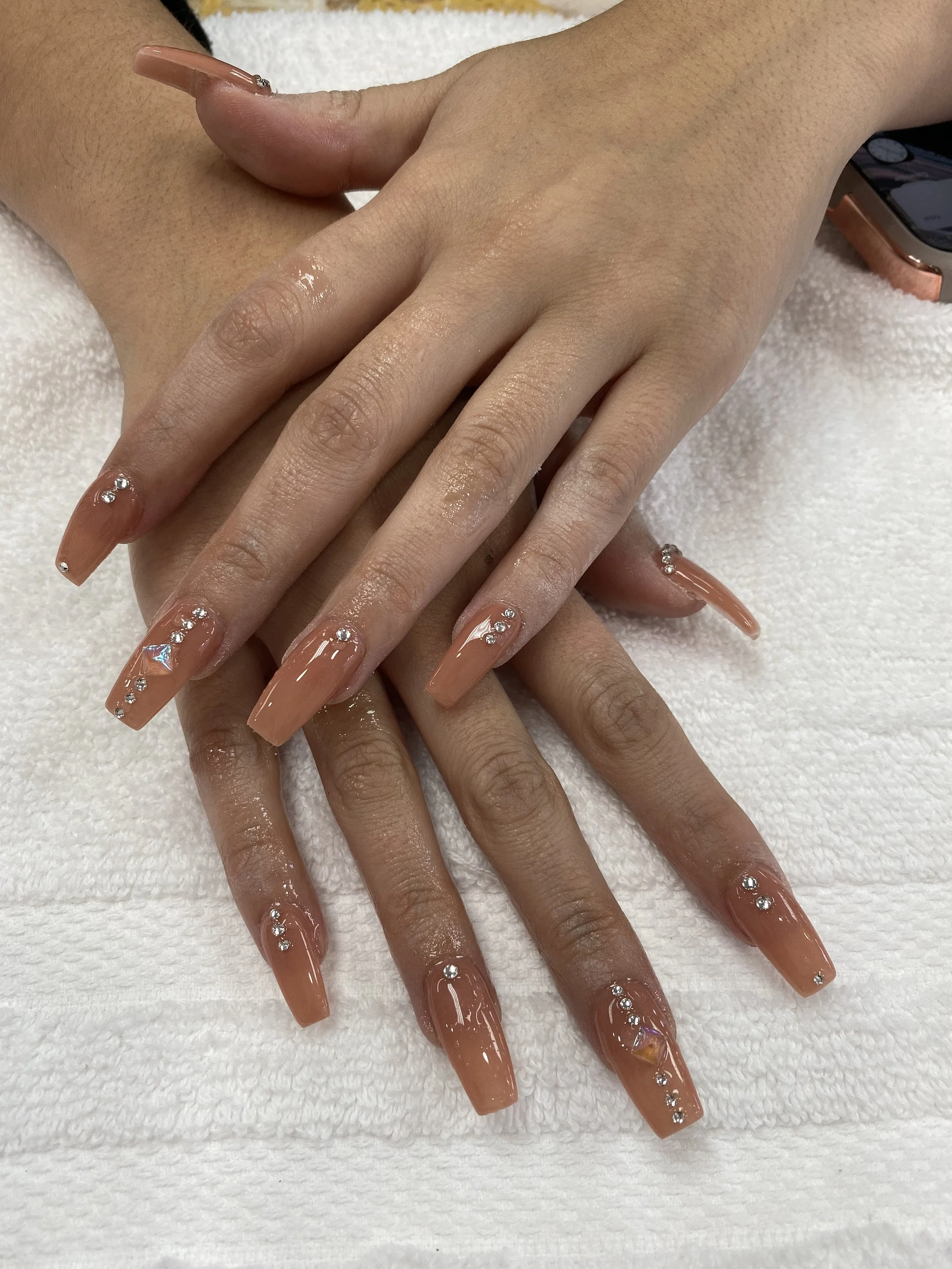 Close-up of hands with long, almond-shaped nude-colored nails decorated with rhinestones and holographic accents. The hands rest on a white towel, and a smartphone is partially visible in the background.