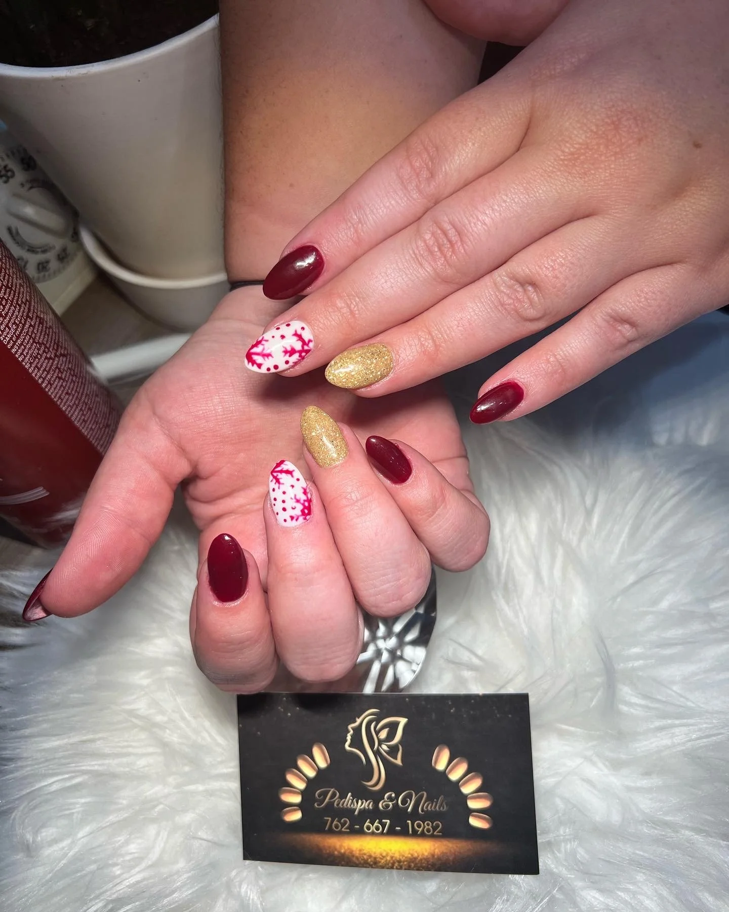 Close-up of a person's hands showing a festive Christmas-themed manicure, with one hand resting on the other. The nails are painted in red, gold, and white with red holiday patterns. In the background, there are nail salon branding and contact inform