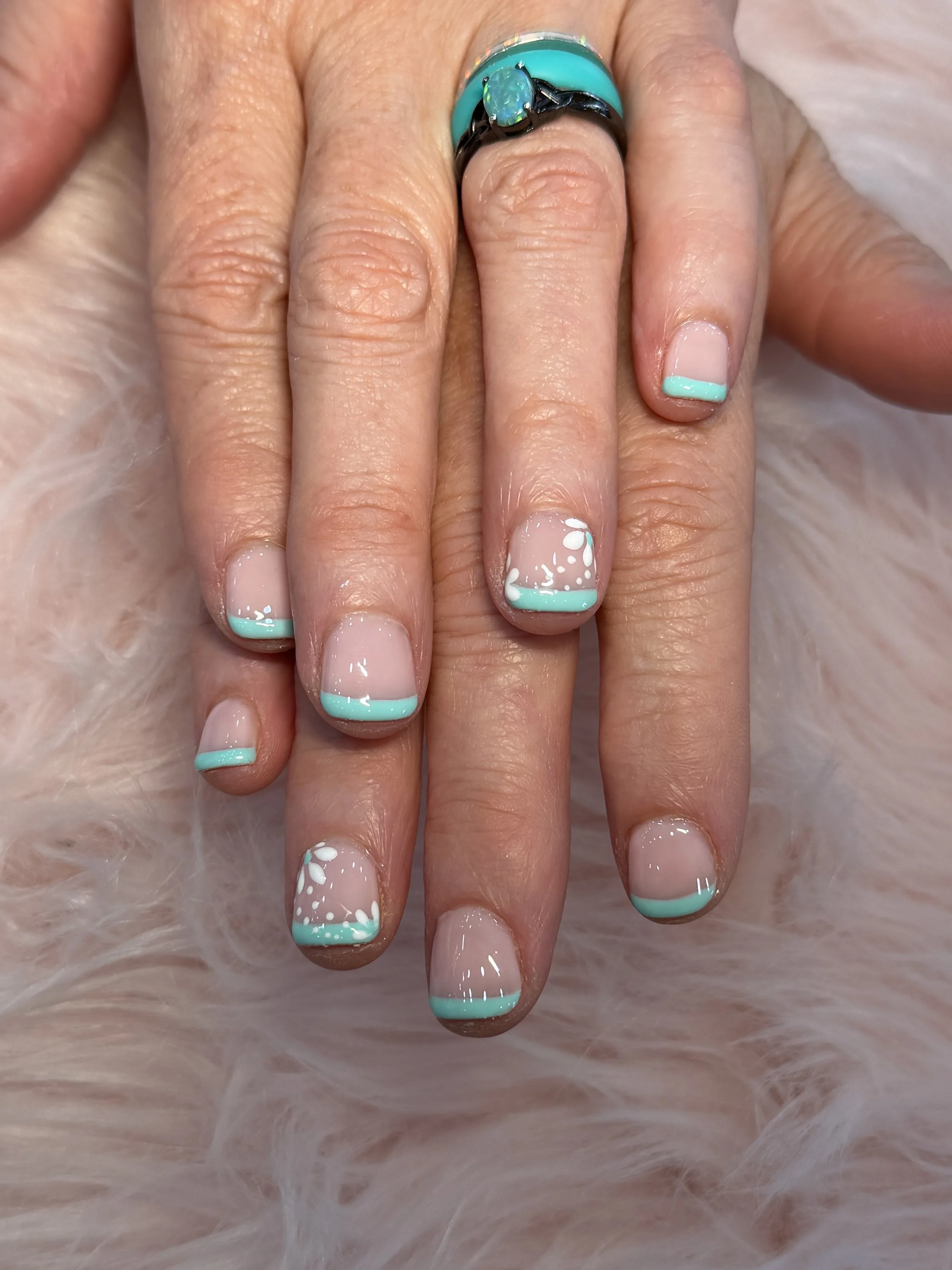 Close-up of a hand with manicured nails, featuring white tips with turquoise accents and floral designs, and a ring with a turquoise stone.
