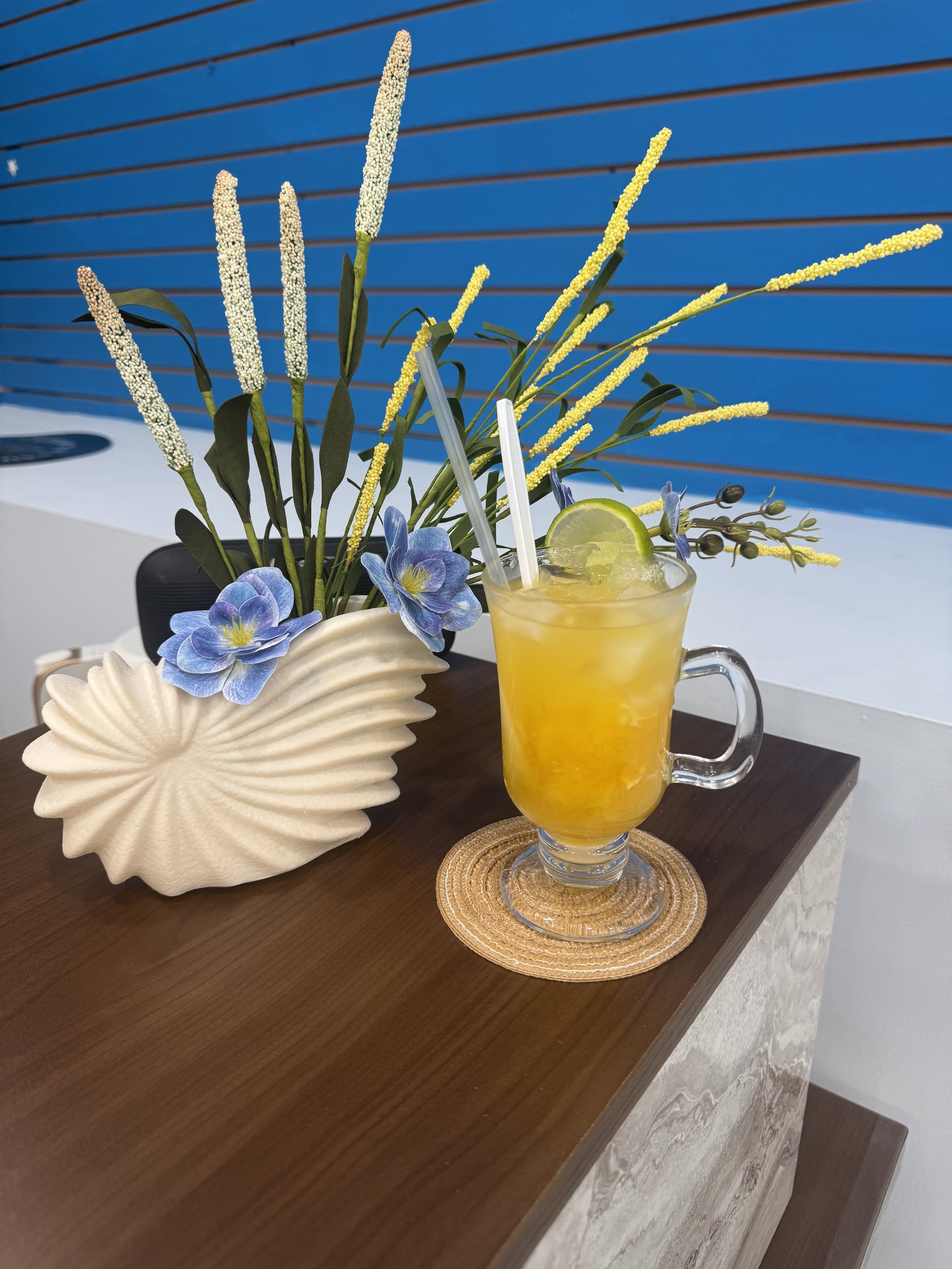 A decorative still life arrangement with a beige shell-shaped vase, artificial blue flowers, a branch with yellow and white filler flowers, and a glass of yellow beverage with lemon, placed on a small woven mat on a wooden table against a blue panele