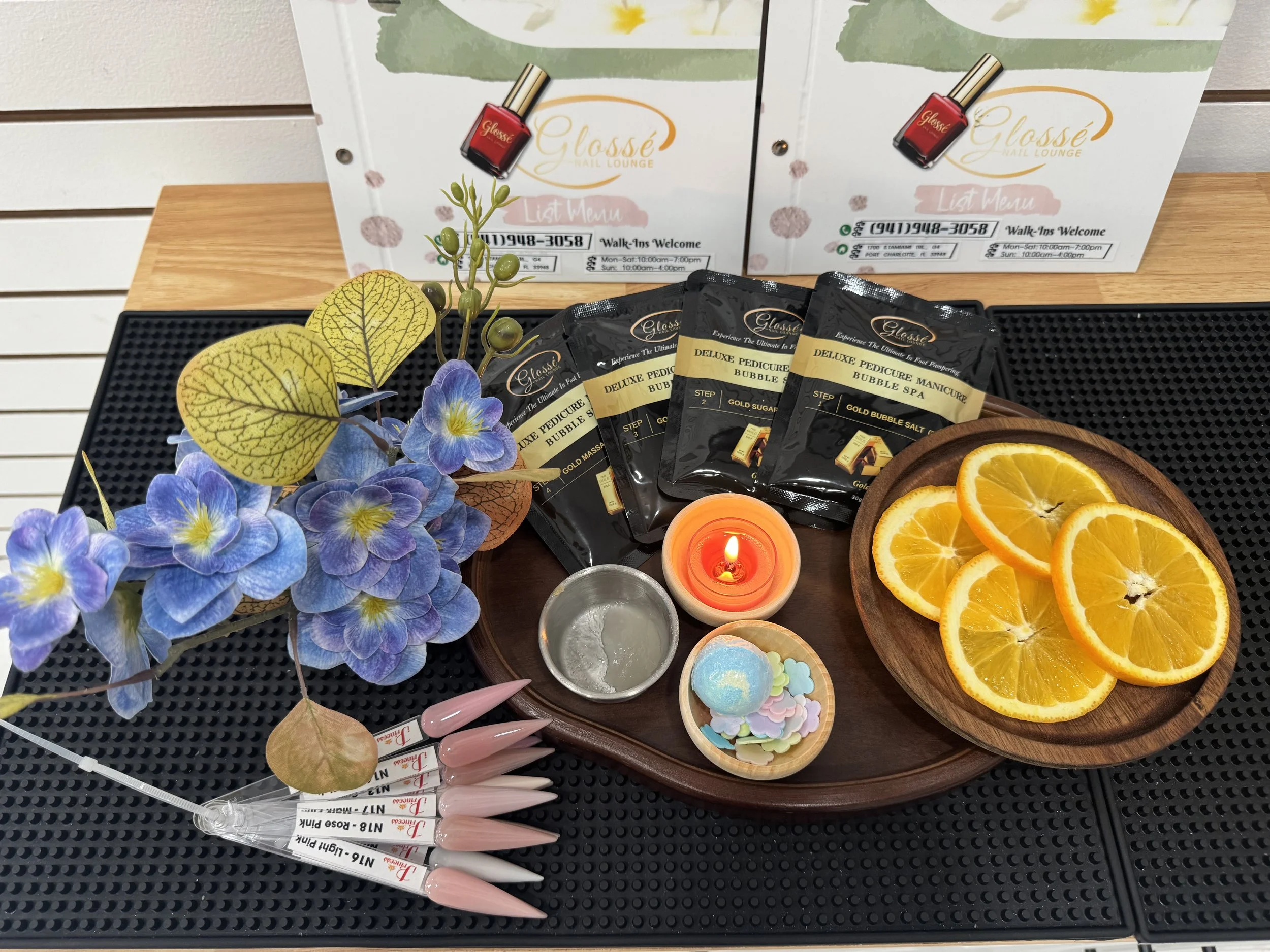 Display of beauty and nail care items including purple and yellow artificial flowers, wrapped pink nail polish brushes, packets of deluxe pedicure manicure bubble spa products, a small orange candle, a wooden plate with sliced oranges, and colorful s