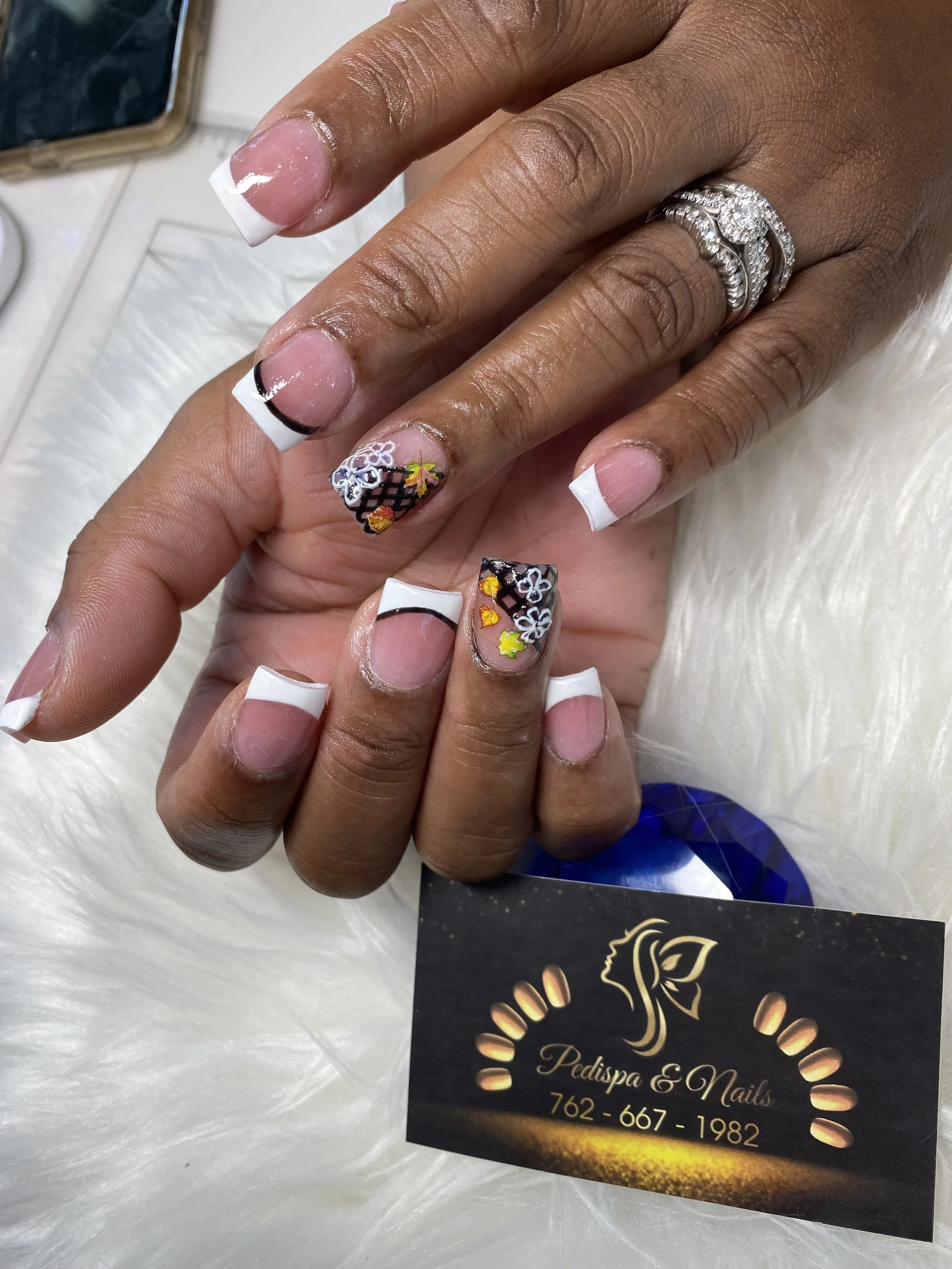 Close-up of a hand with manicured nails, some painted with French tips and others with black base and colorful floral designs. The hand is wearing multiple silver rings with embedded stones. There is a black business card from Pedispa & Nails visible