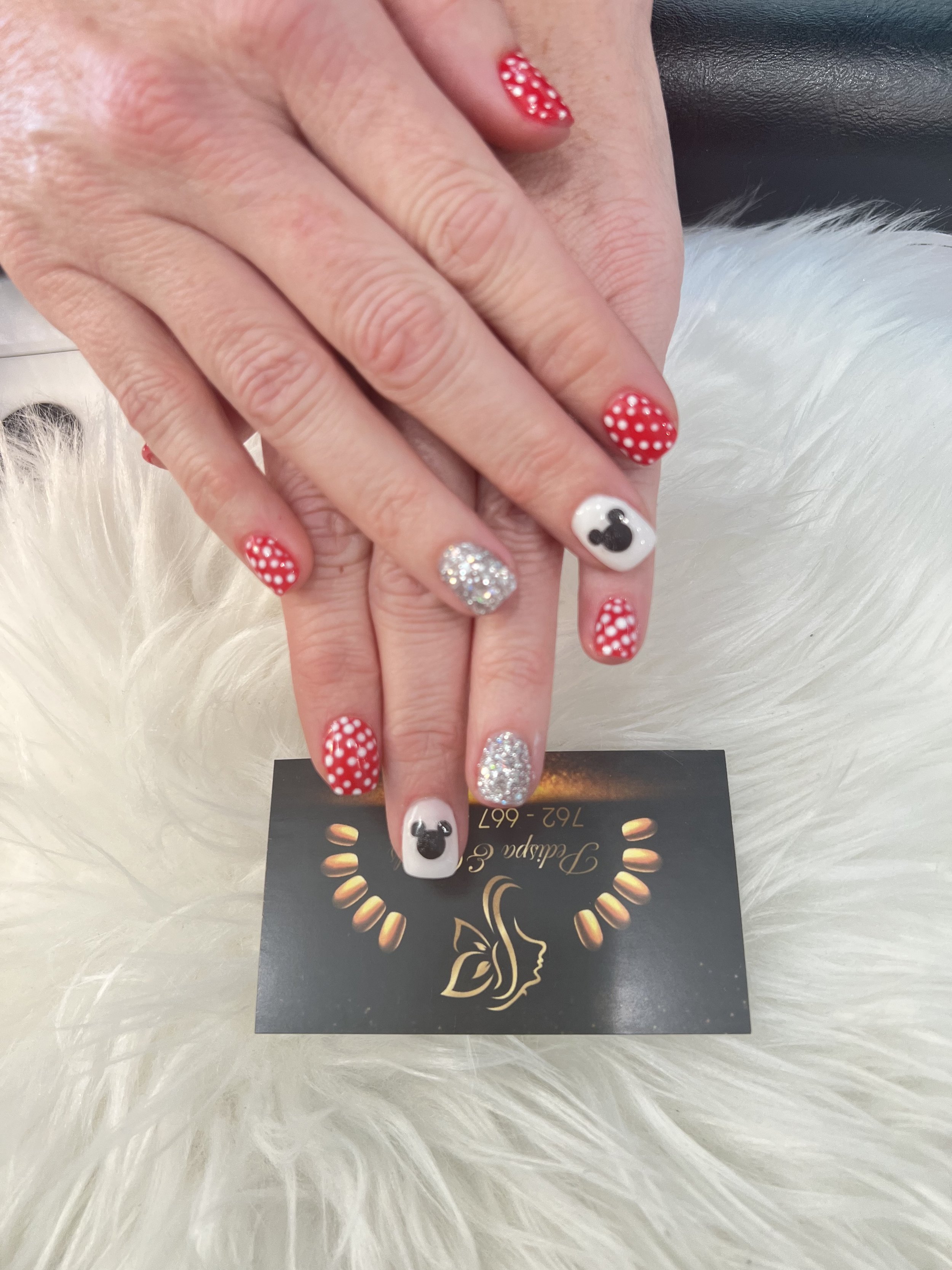 Close-up of a person's hands resting on a white furry surface, showing holiday-themed decorated nails, with one nail featuring a Mickey Mouse silhouette. The hands are held over a black card with gold and white lettering and a butterfly logo.