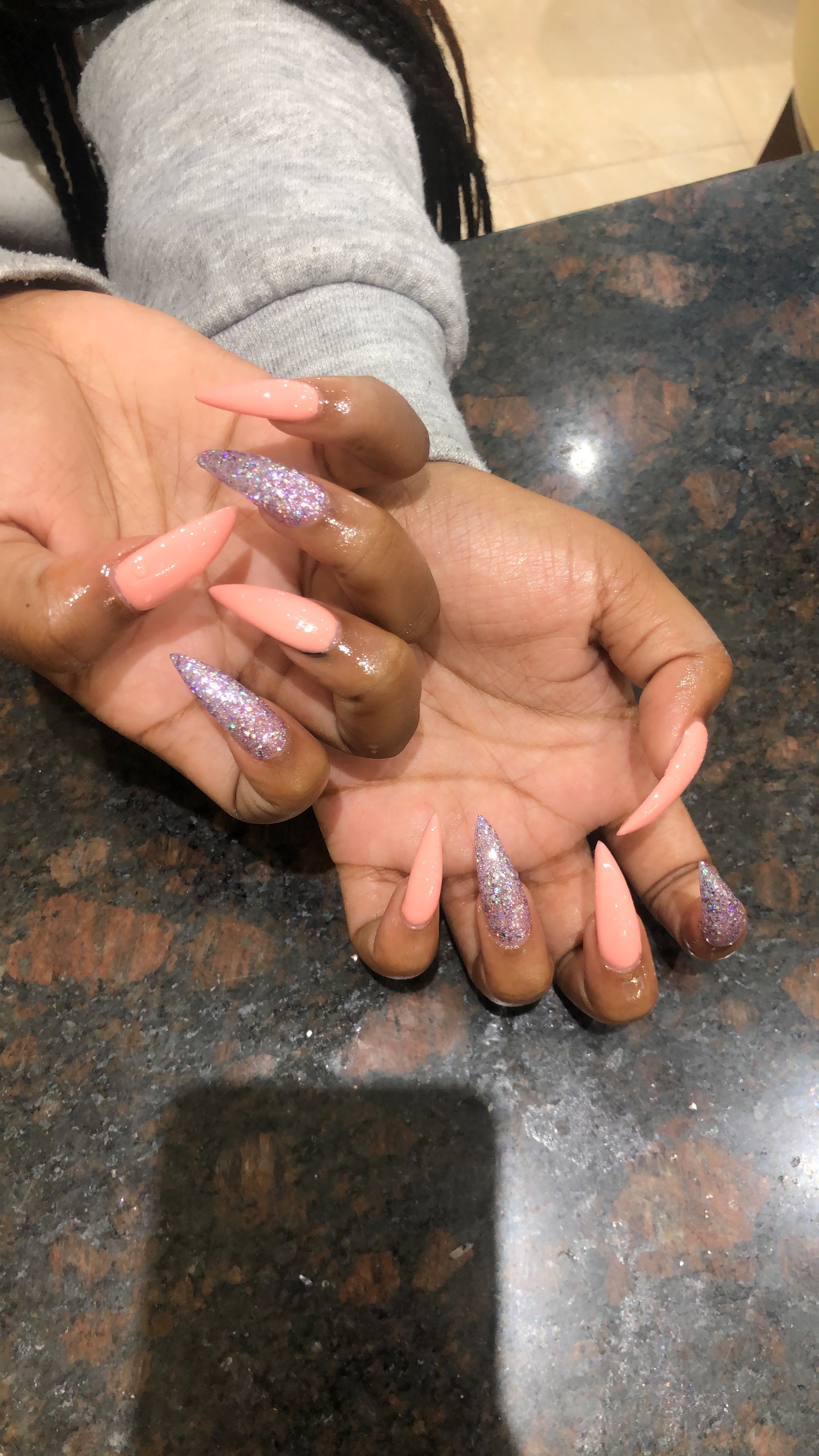 Close-up of a person's hand with long, almond-shaped, peach-colored nails and glittery accent nails, resting on a dark, speckled granite countertop.