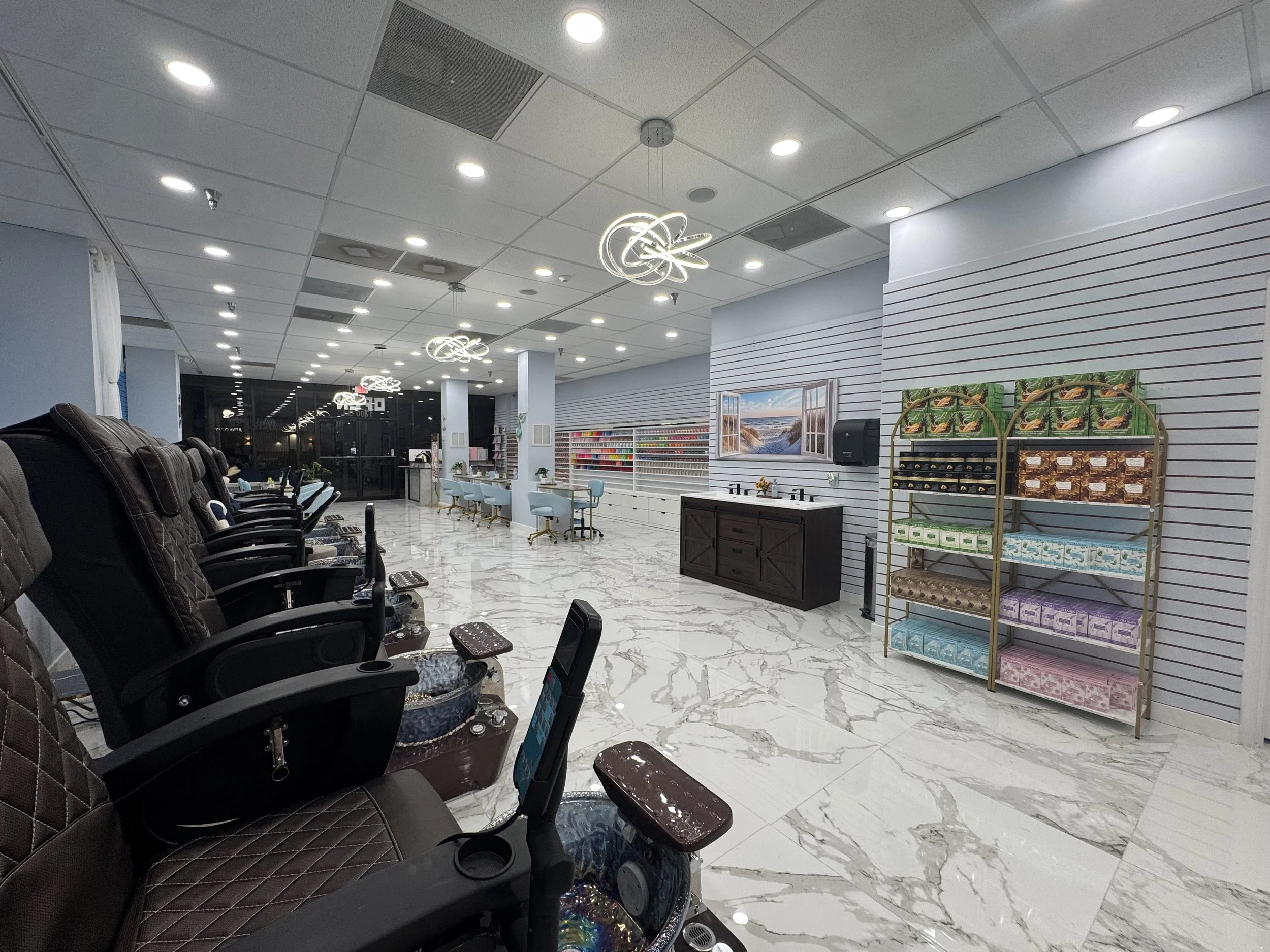 A salon interior with multiple massage chairs along the left, white and marble floors, a reception desk in the back, shelves with colorful products, wall art of a beach scene, and decorative ceiling lights.