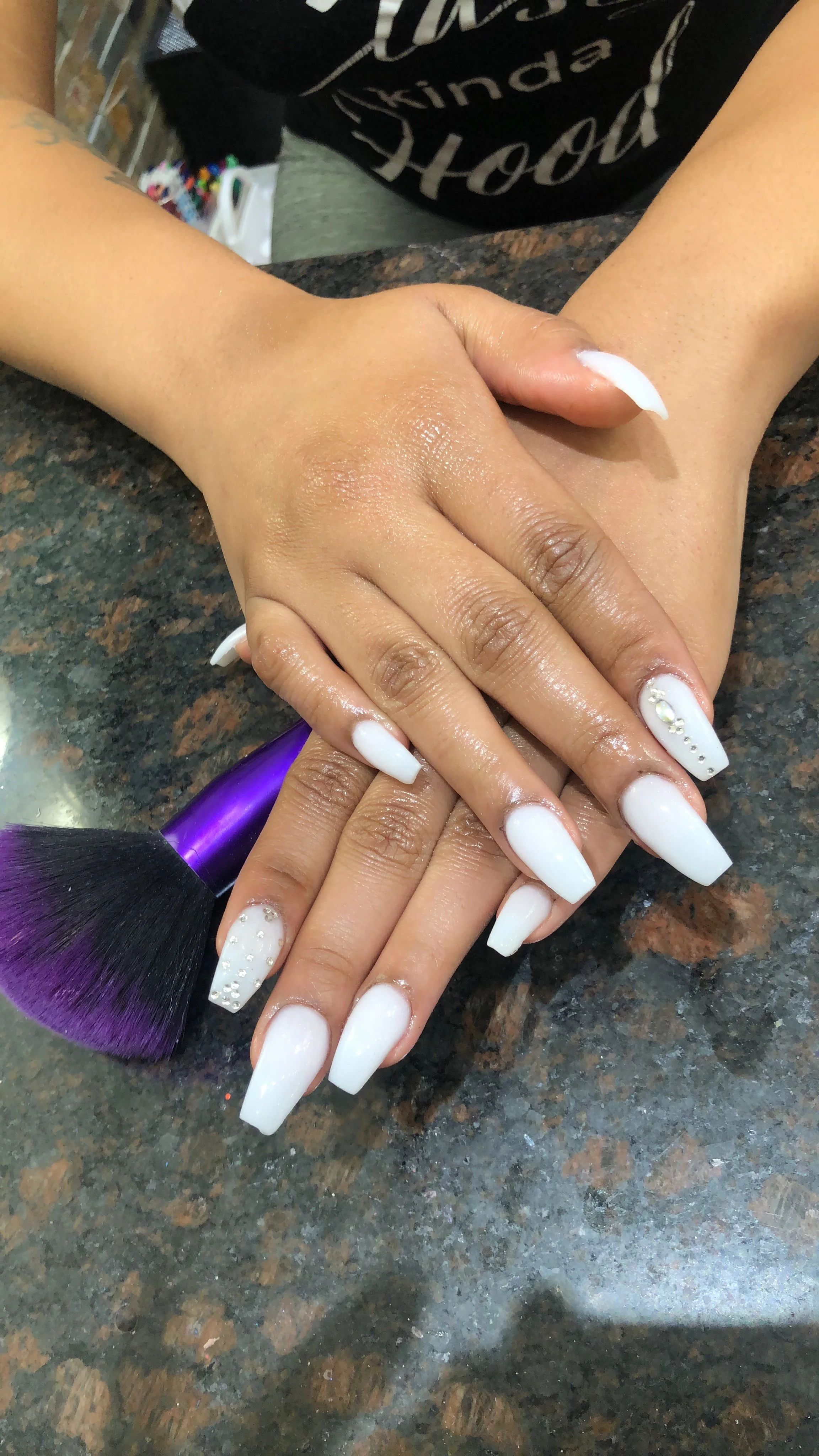 Hands with white acrylic nails and rhinestone embellishments, one hand resting on top of another, on a granite countertop, with a makeup brush visible.