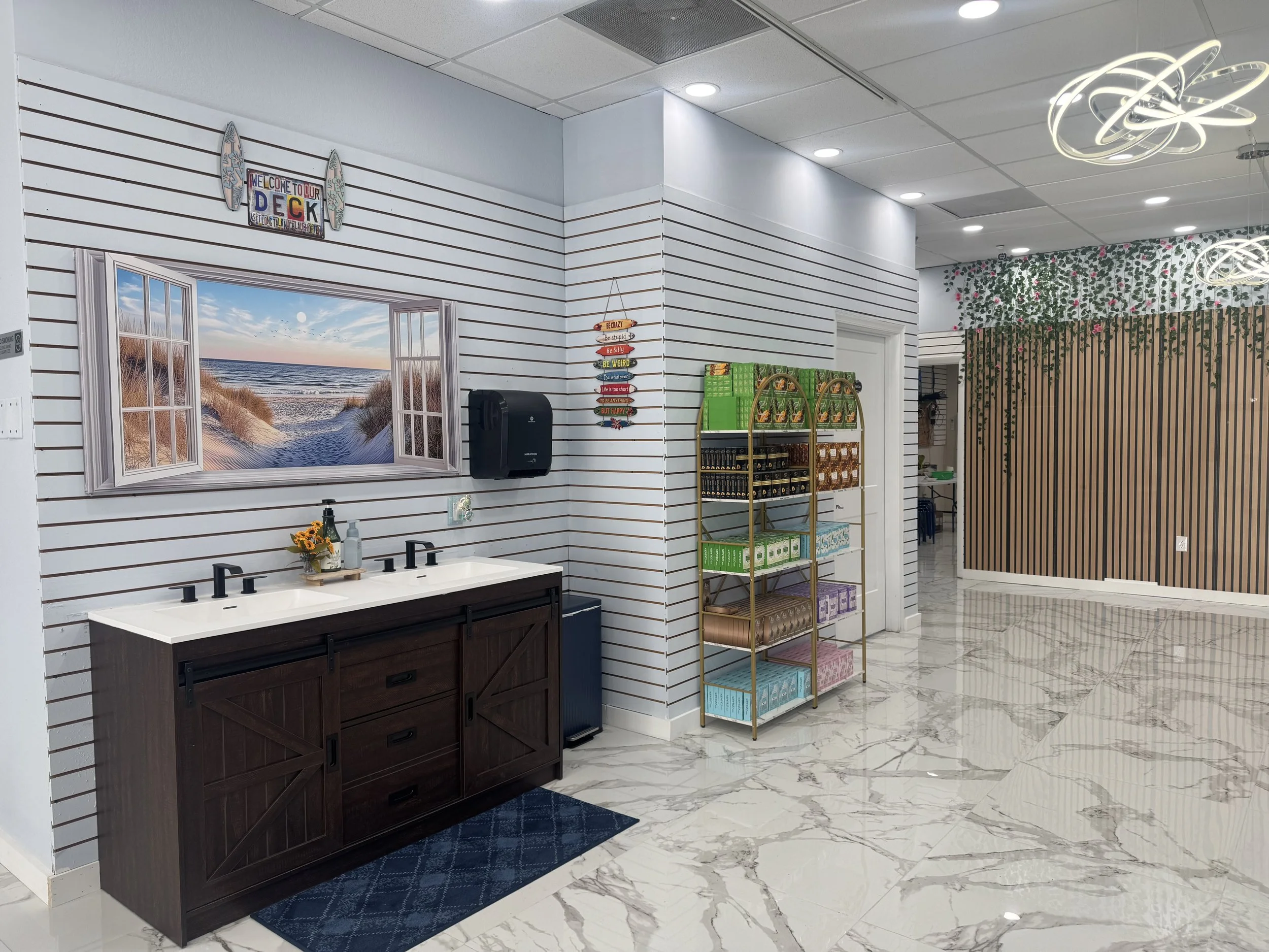 Interior of a store with white shiplap walls, a mural window view of a beach, a dark brown vanity with three sinks, a gold shelf stocked with green, purple, and orange packages, a wall sign that reads 'Welcome to our deck,' and decorative elements in