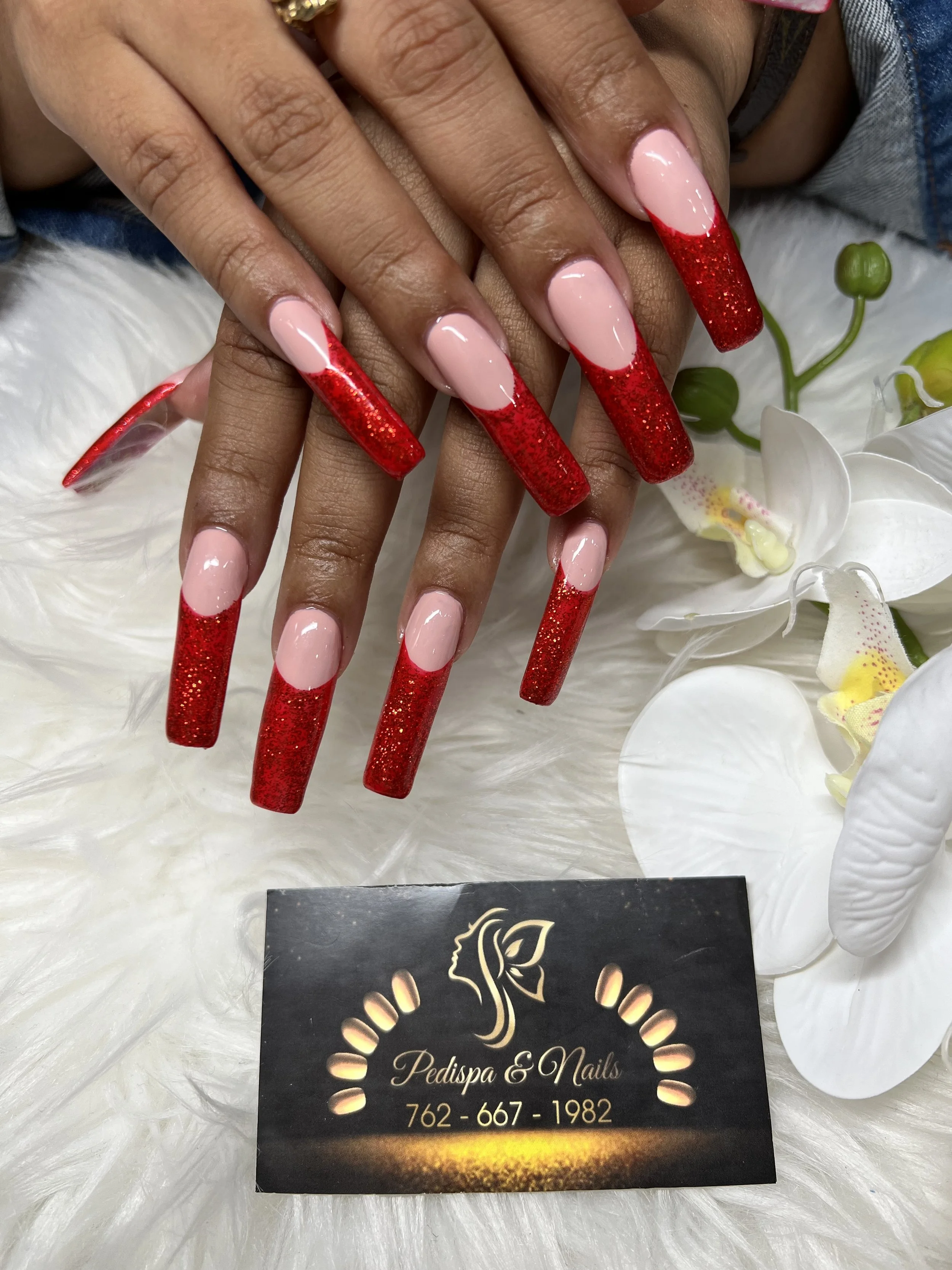 Close-up of a woman's well-manicured hands with long, almond-shaped nails painted in a soft pink with red glittery tips. The hands are resting on a white furry surface with some white orchid flowers nearby. A business card for Pedispa & Nails, with s