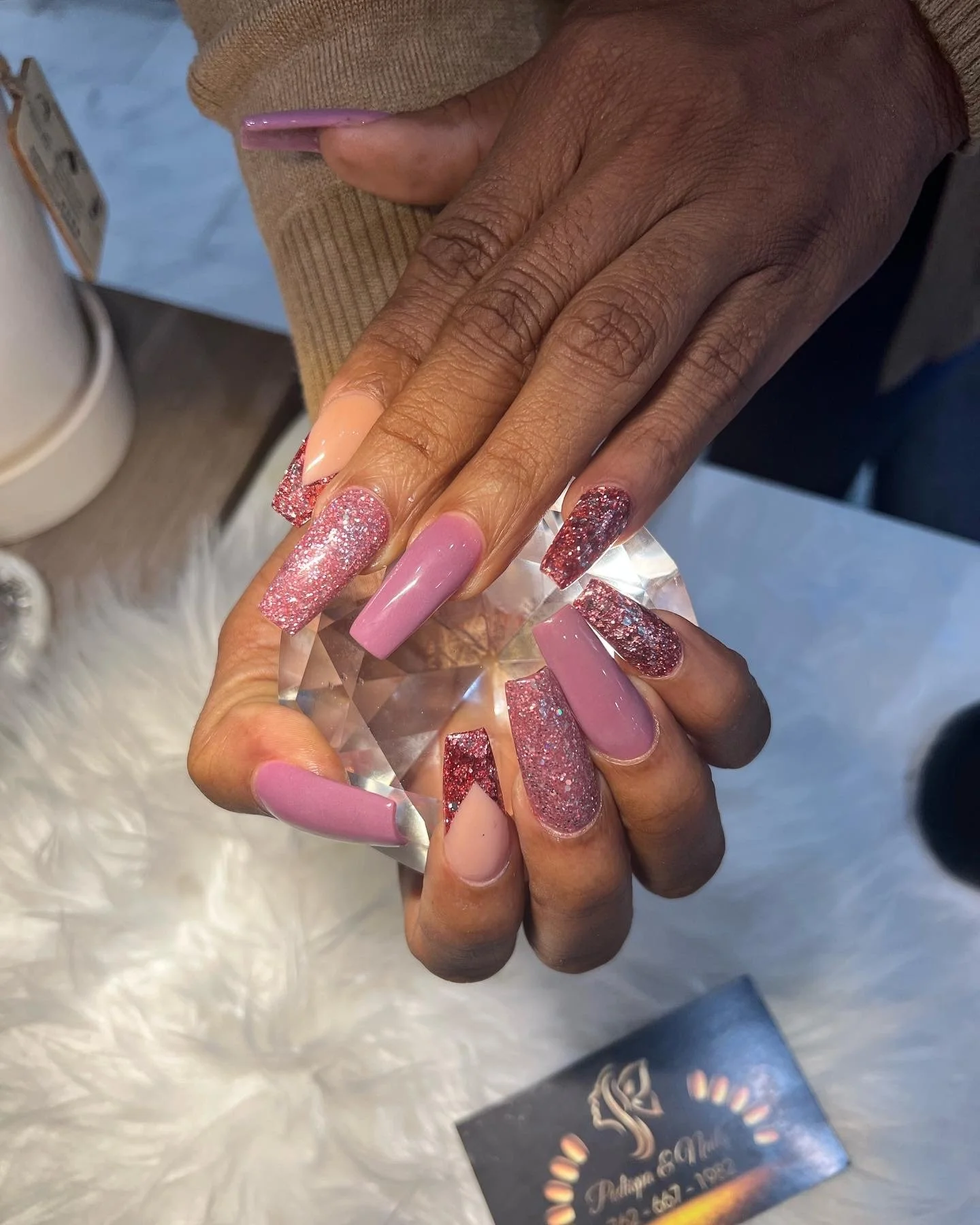 Close-up of a person with dark skin showing off their freshly painted and decorated long acrylic nails, which are pink with glitter accents in a salon or nail spa setting.