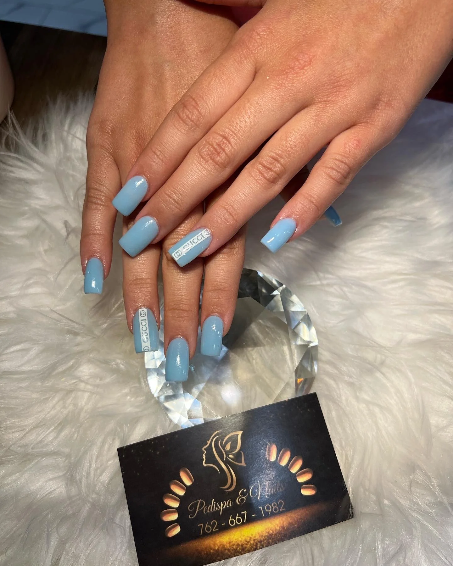 Close-up of a woman's hands with light blue acrylic nails, one nail displaying the Gucci logo. The hands rest on a white, furry surface, with a black business card for Pedispa & Nails underneath, showing a logo, contact number, and establishment year