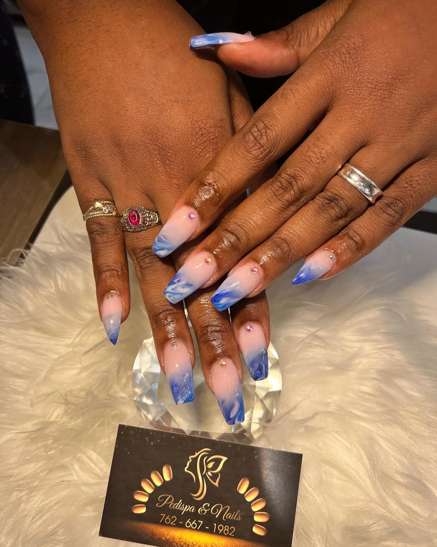 Close-up of a person’s hands with a detailed blue and white marble nail design, adorned with small rhinestones near the cuticles, showcasing jewelry including rings, with a business card for Pedispa & Nails visible at the bottom.