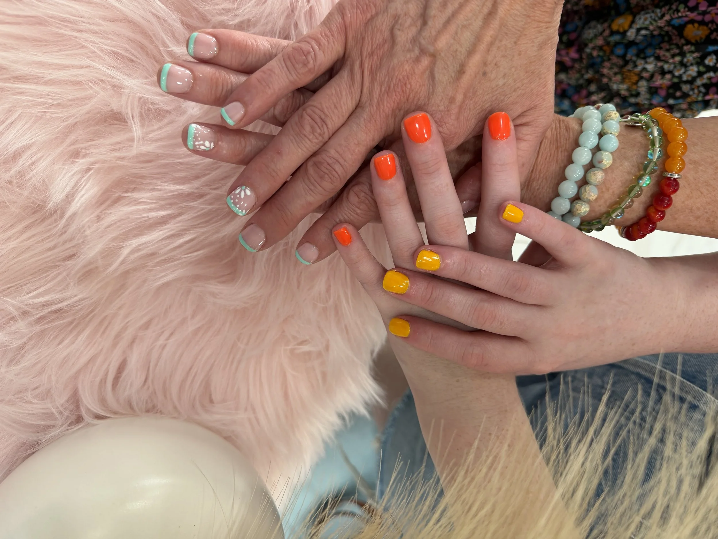 Close-up of hands with manicured nails, the larger hand has natural nails with a mint green tip and white floral design, the smaller hand has bright orange and yellow nails. The hands are resting on pink faux fur, with beaded jewelry visible on the w