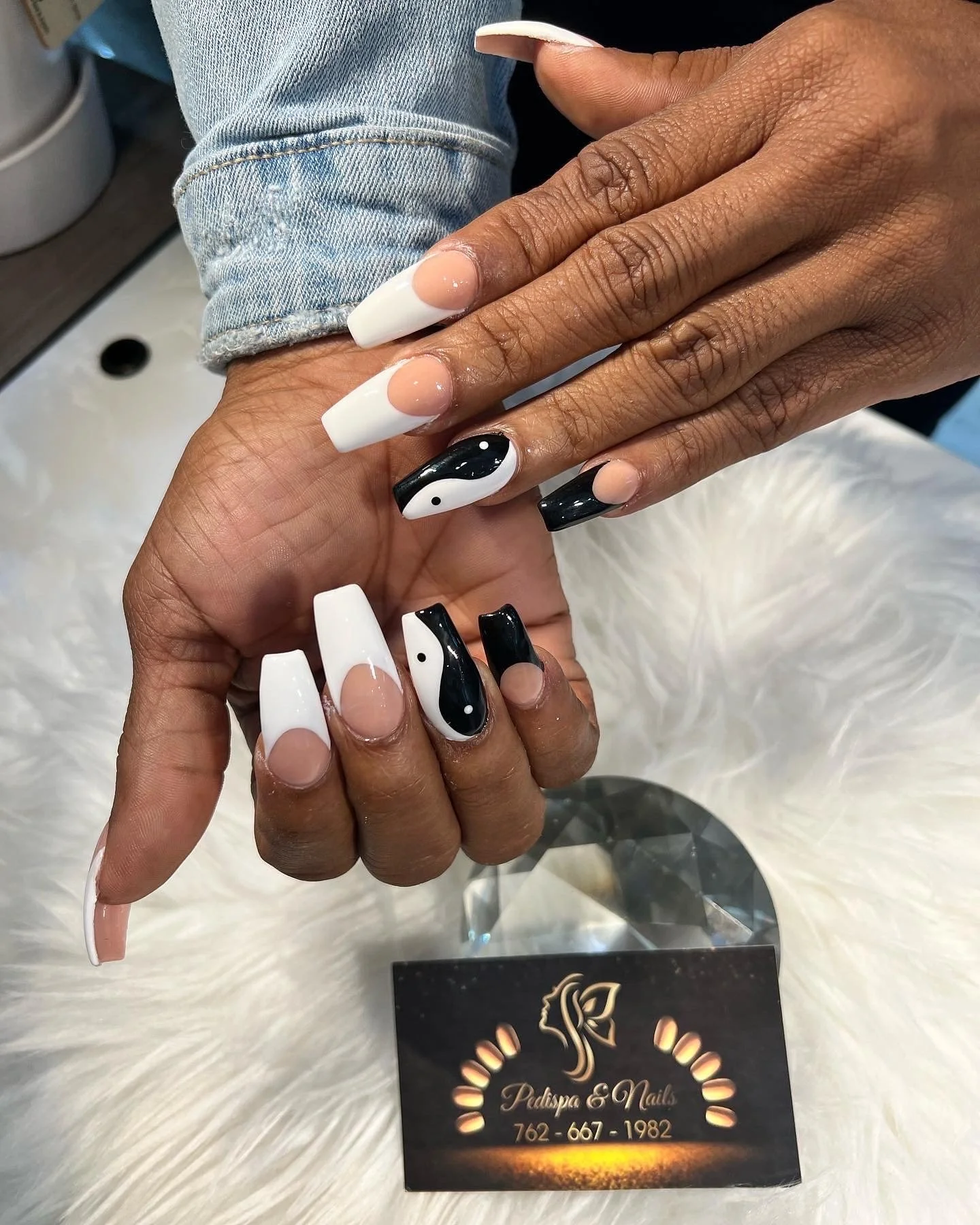 Close-up of two hands with decorated acrylic nails, one with black and white yin-yang design, and the other with a white and nude design. The hands are resting on a white, fur-like surface, with a business card that reads 'Padispa & Nails' with a pho