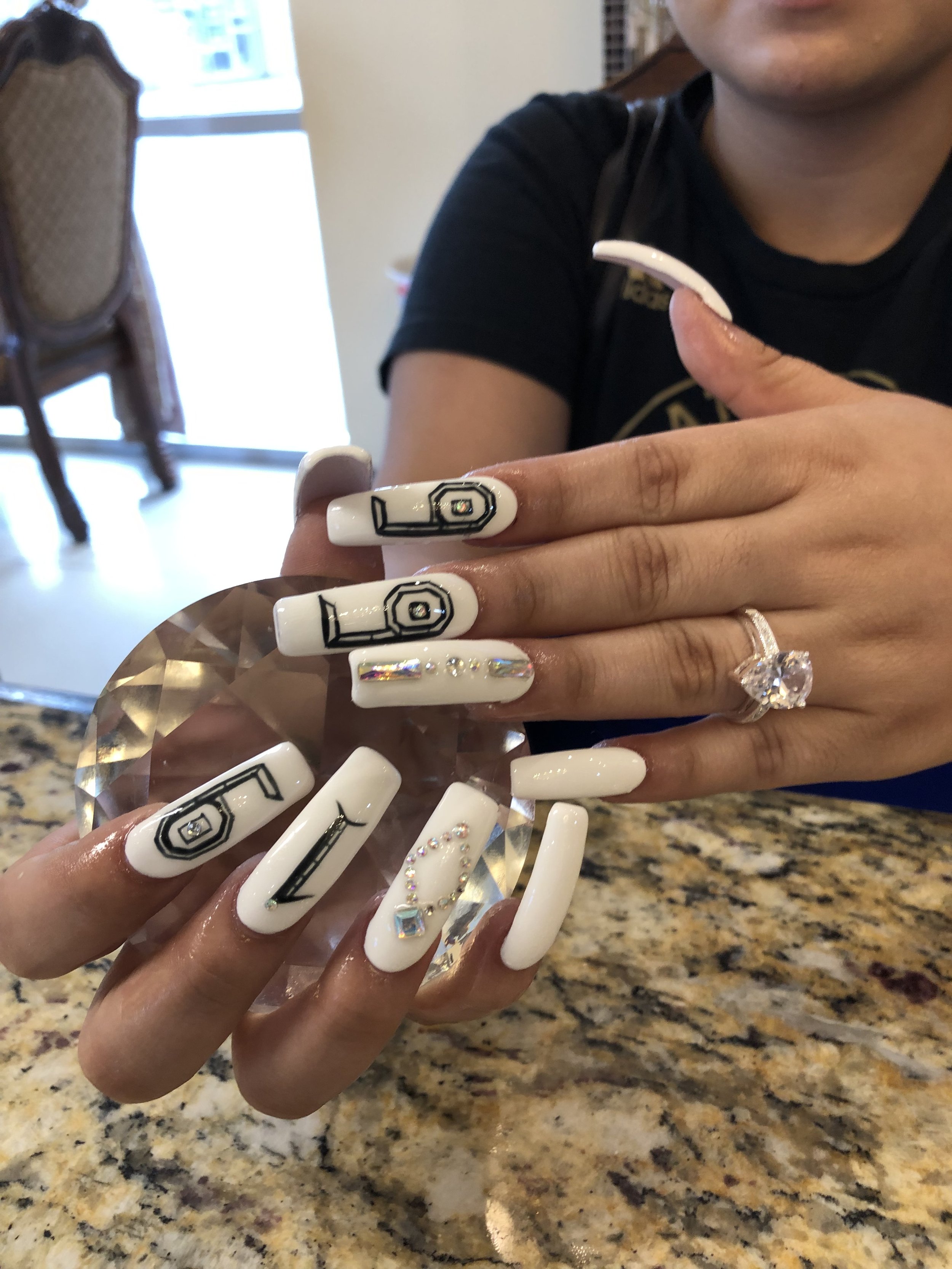 Close-up of a woman's hand with white, decorated nails, holding a large crystal, and wearing a ring with a large heart-shaped stone. The background shows a countertop, a person's chin and part of their face, and some furniture.