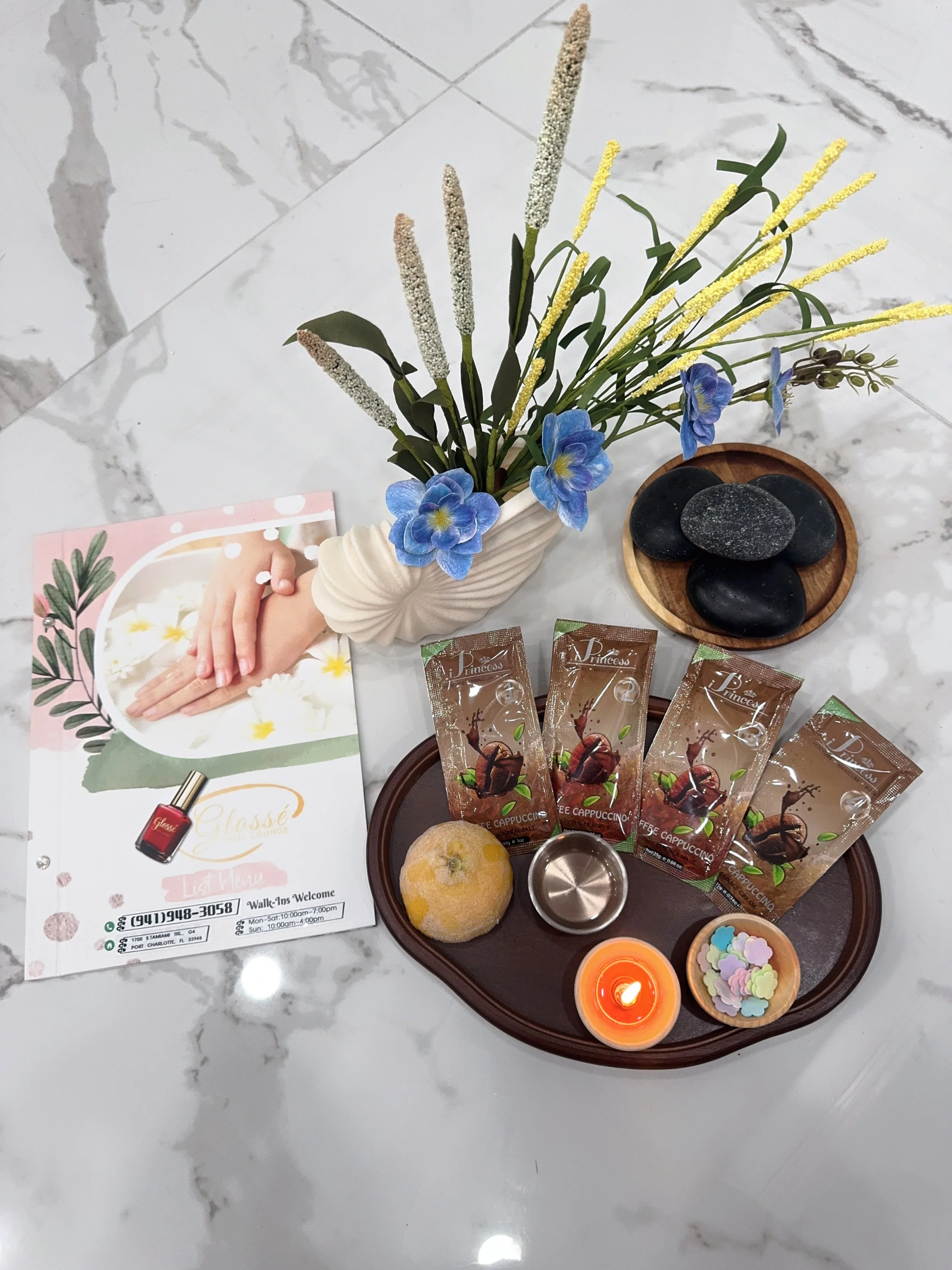 A marble table with a decorative flower arrangement in a white vase, containing yellow, white, and blue flowers, alongside a wooden tray with black stones, a small orange candle, a small bowl of pastel-colored confetti, coffee sachets labeled 'Coffee Cappuccino,' a menu flyer, a bread roll, and a small metallic container.