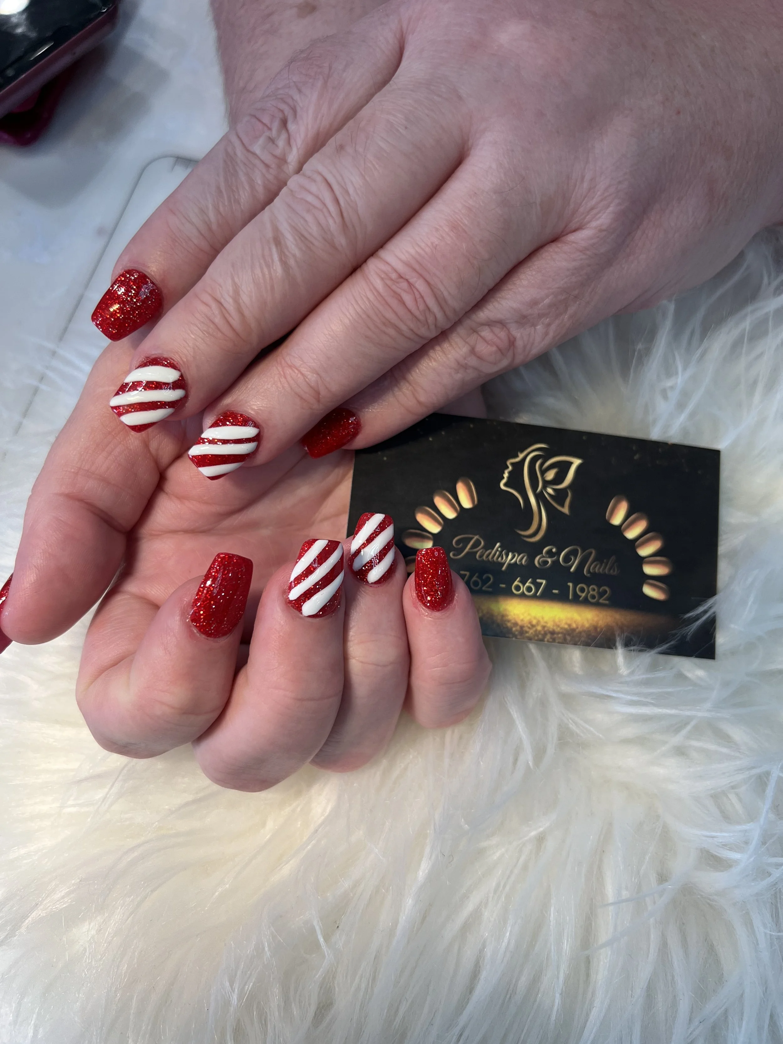 Close-up of a hand with painted nails, featuring red glitter and white and red striped nail art, resting on a soft, white, fluffy surface with a business card partially visible underneath.