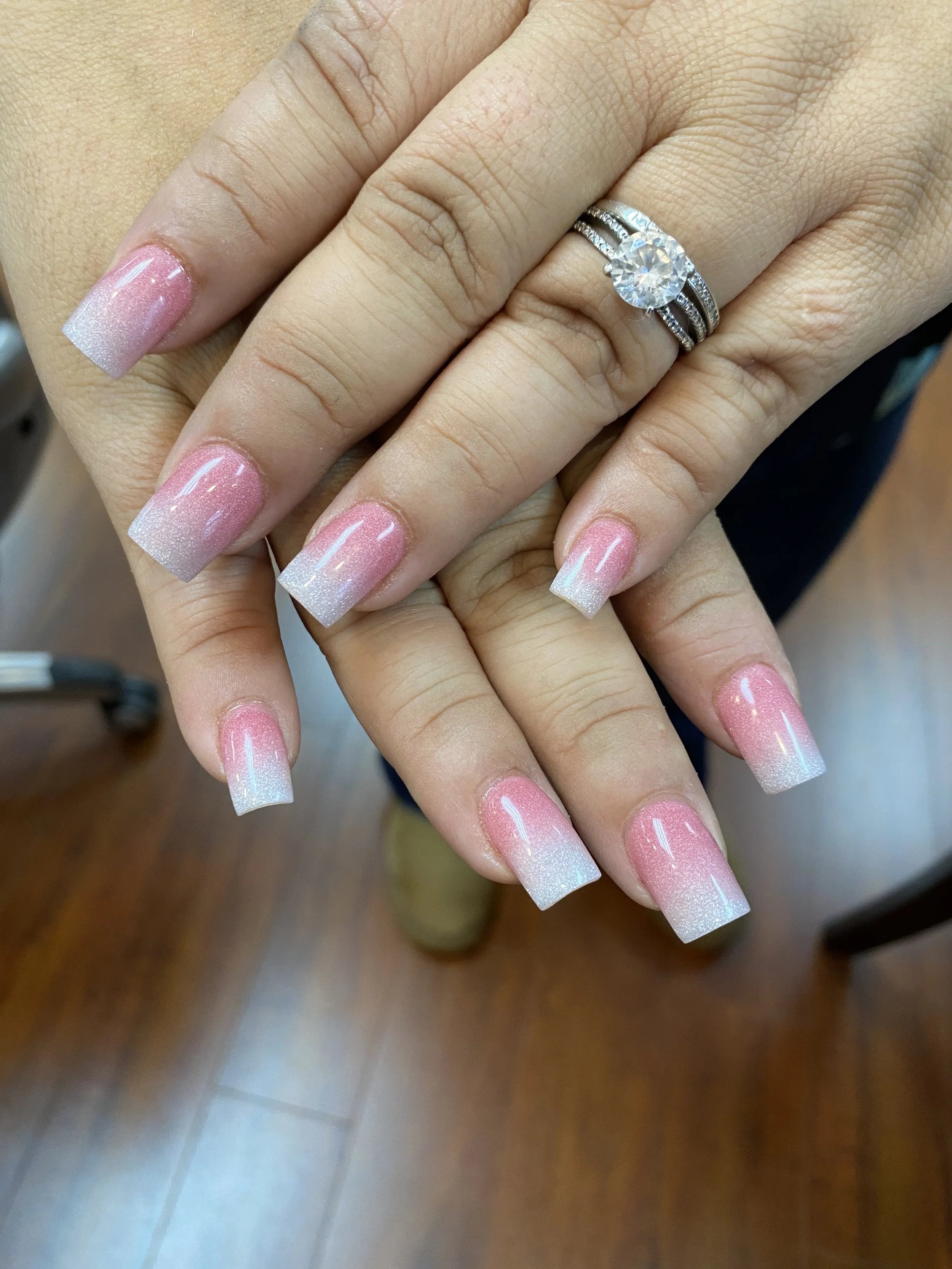 Close-up of a person's hand showing manicured nails with French tips and a ring with a large round diamond surrounded by smaller diamonds.