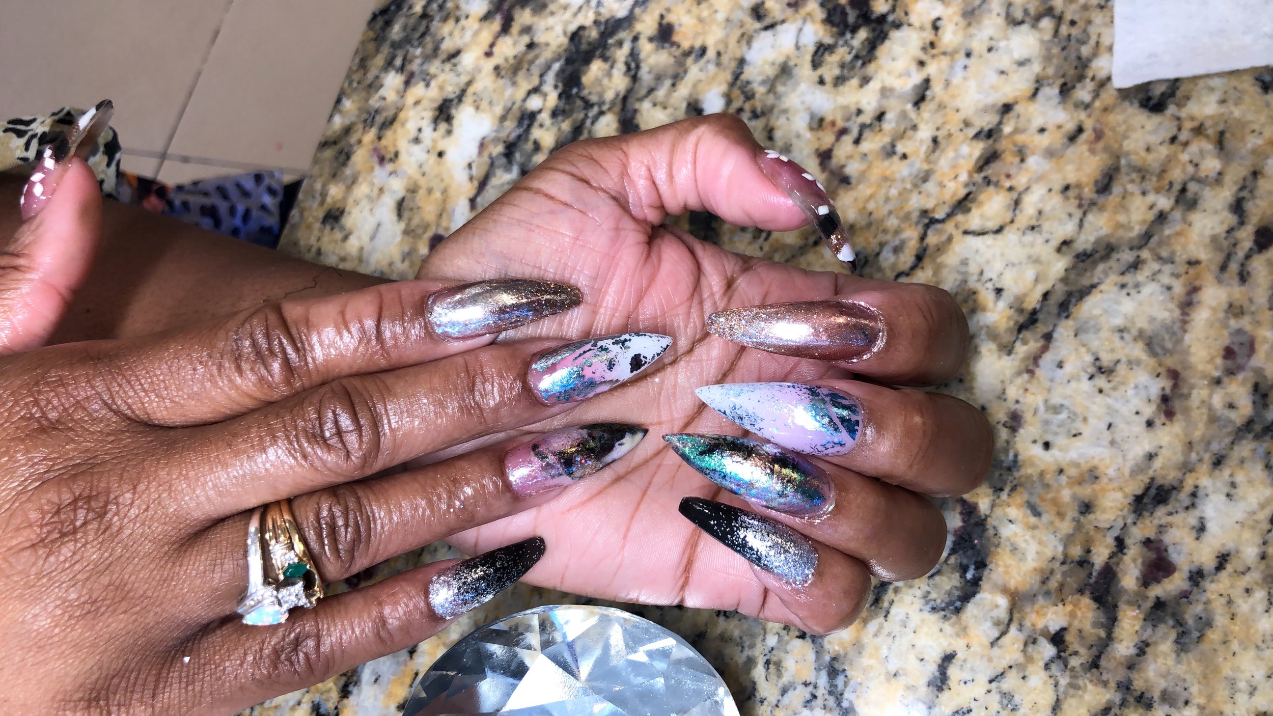 A person with dark skin showing off long, pointed, multi-colored, holographic stiletto nails with a mix of shiny, glitter, and marble-like designs, resting on a granite countertop.