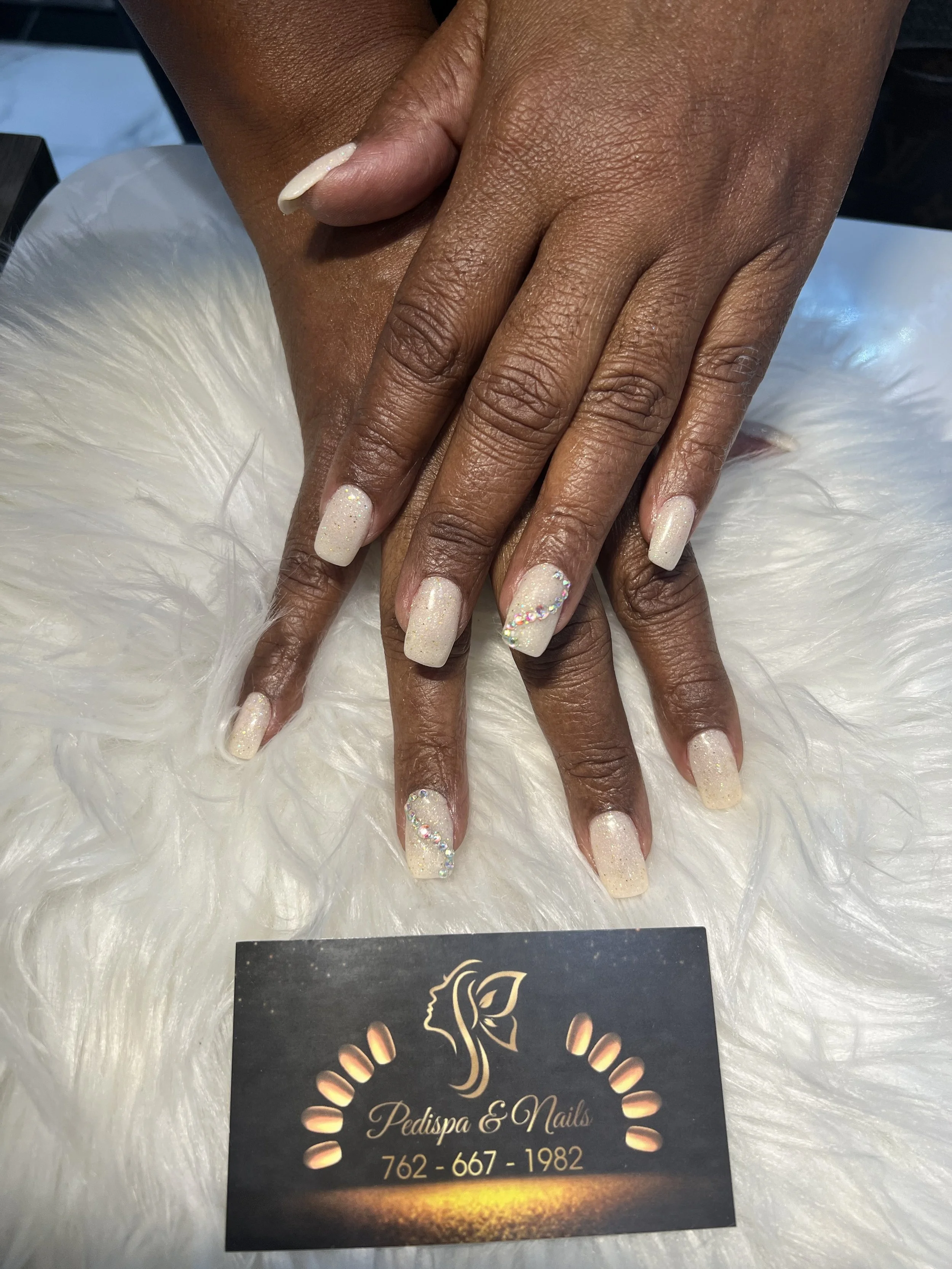 Close-up of a person's hands with manicured nails resting on a white furry surface. The nails are painted in a glittery, pale color with rhinestones on some nails. A black business card with gold and white text is partially visible, displaying the na