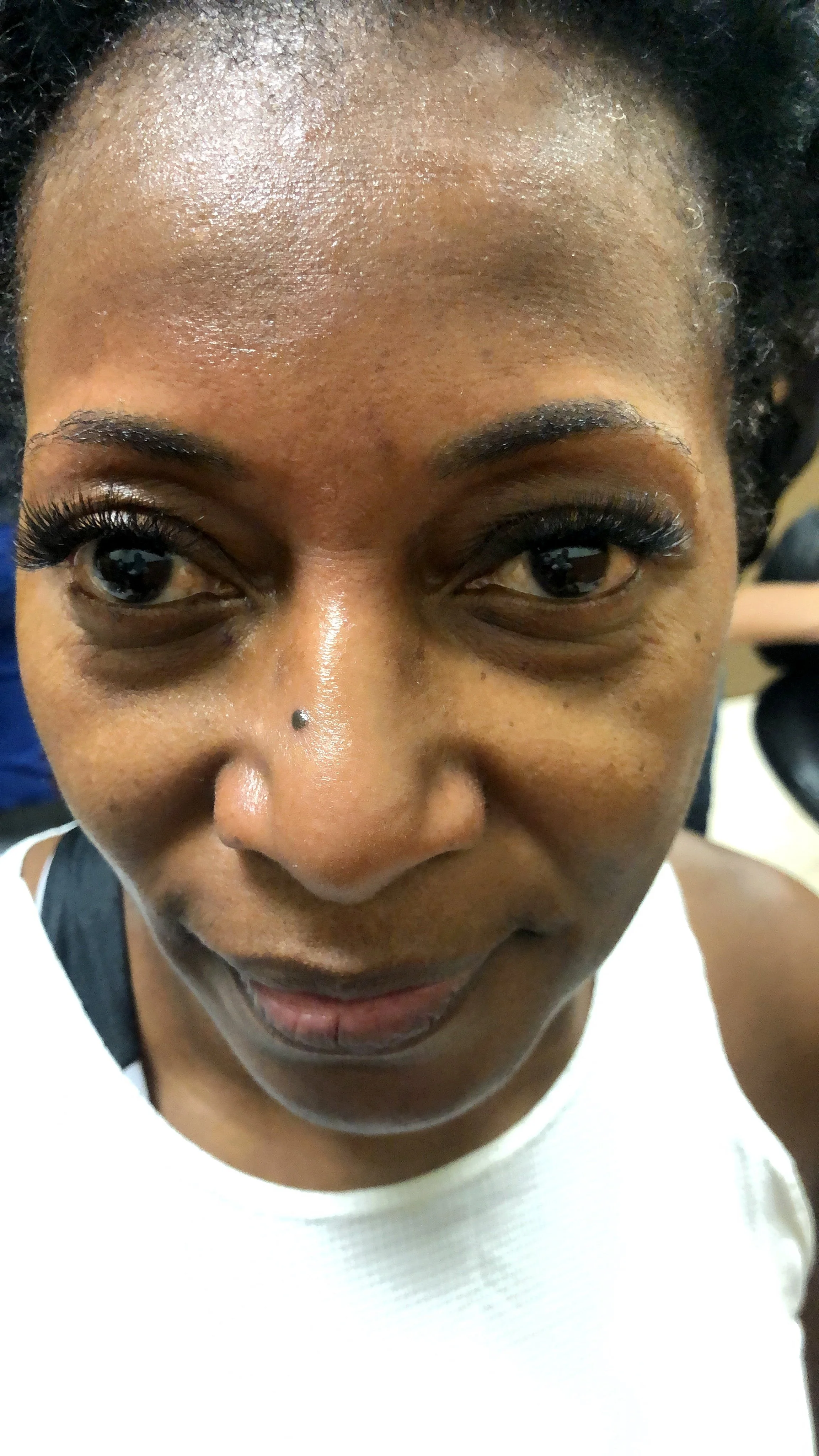 Close-up of a woman with dark skin, black hair, and makeup, in a white top, looking at the camera.