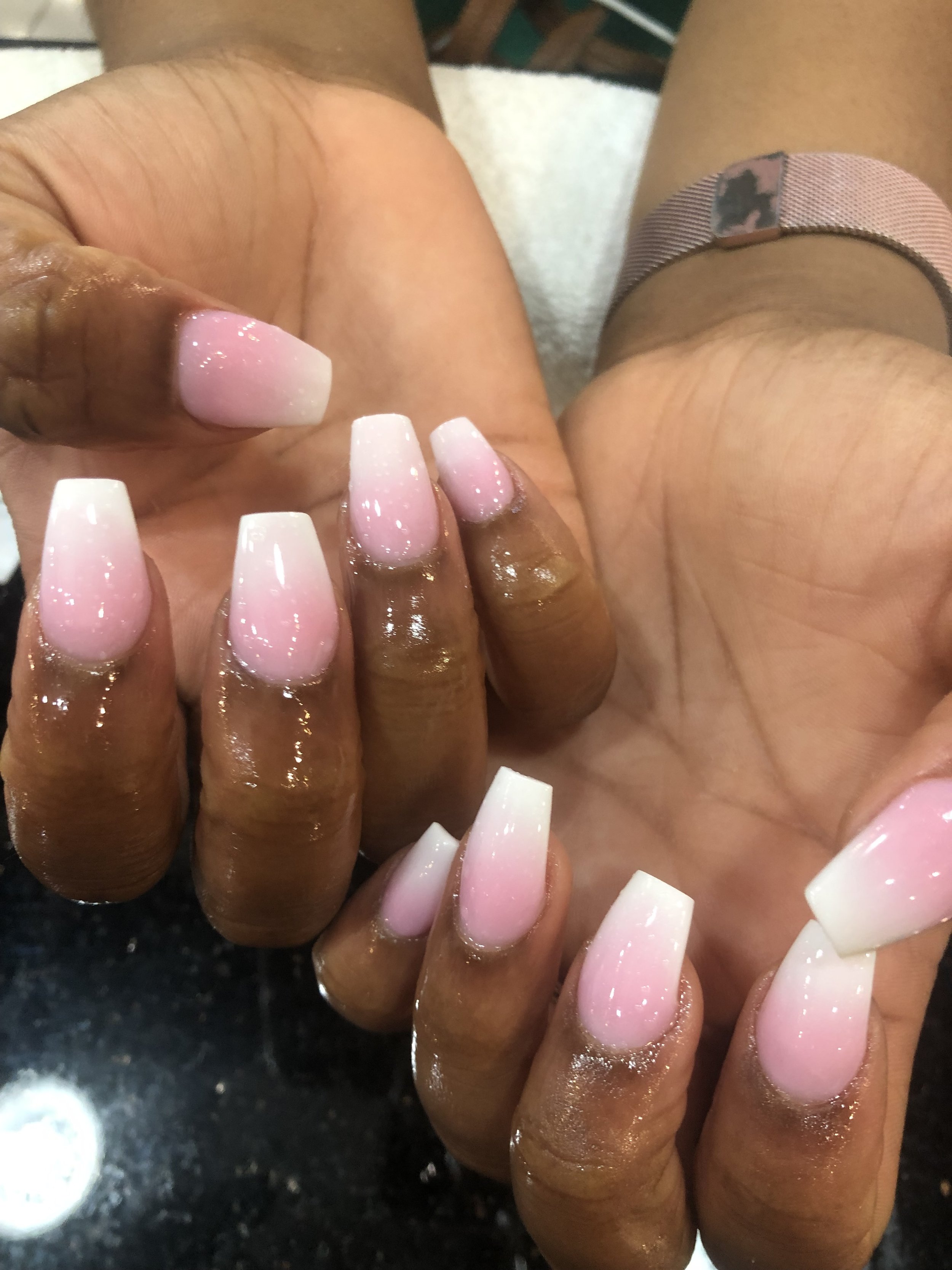 Close-up of manicured nails with an ombre pink and white gradient, held by female hands with brown skin, on a dark surface.