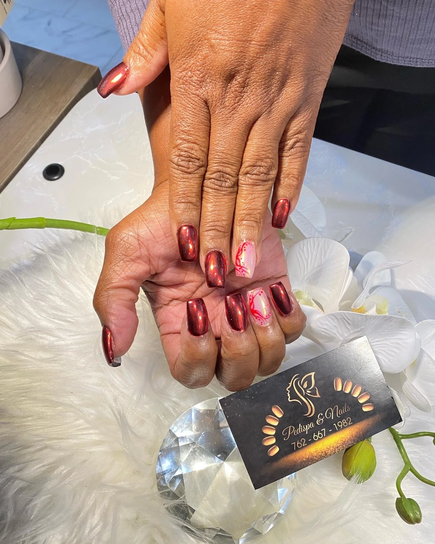 A person's hand with a metallic red manicure, holding a hand with white and red-detailed nails, over a white fur surface with orchids and a business card for Pedispa & Nails.