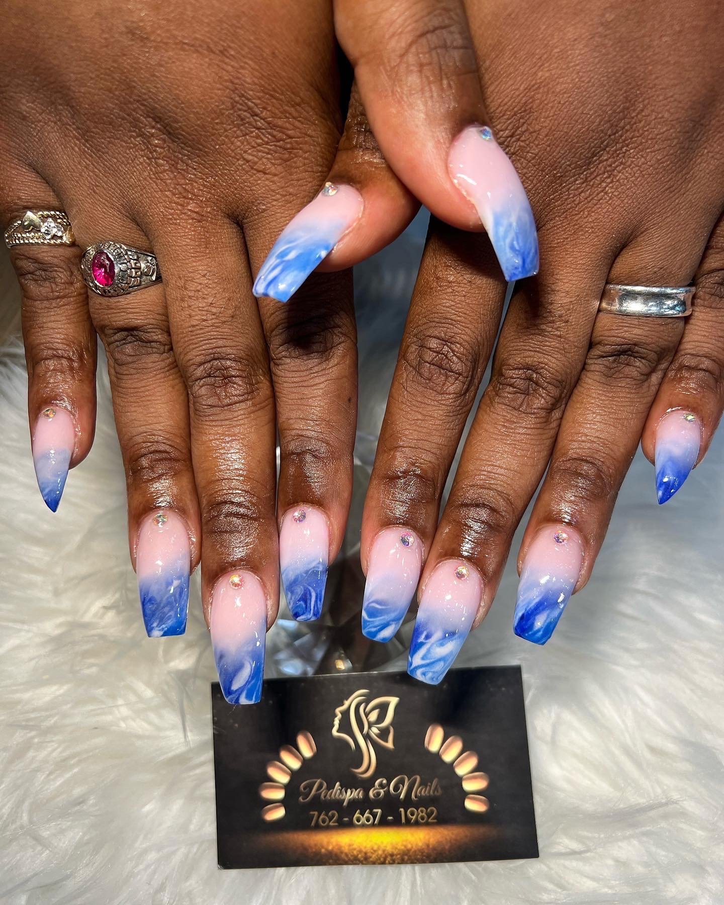Close-up of hands with gel manicured nails featuring a blue and white swirl design, wearing rings, with a business card from a nail salon in the background.