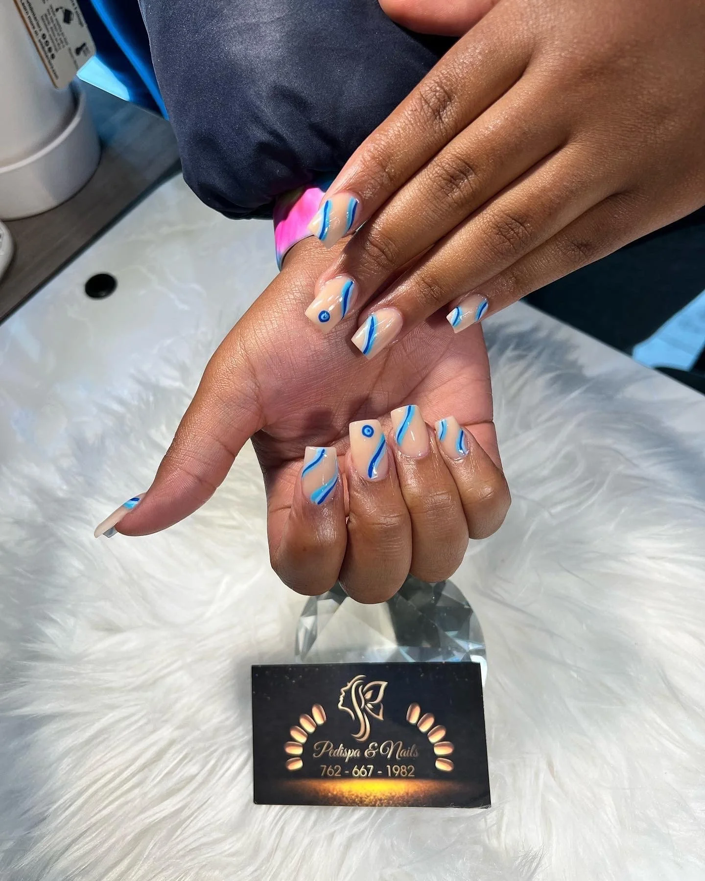 Close-up of a person's hand with nail art featuring a swirl design in beige, blue, and white, placed on top of another hand with similar nails. A business card for Pedspa & Nails is visible underneath showing contact number 762-667-1982.