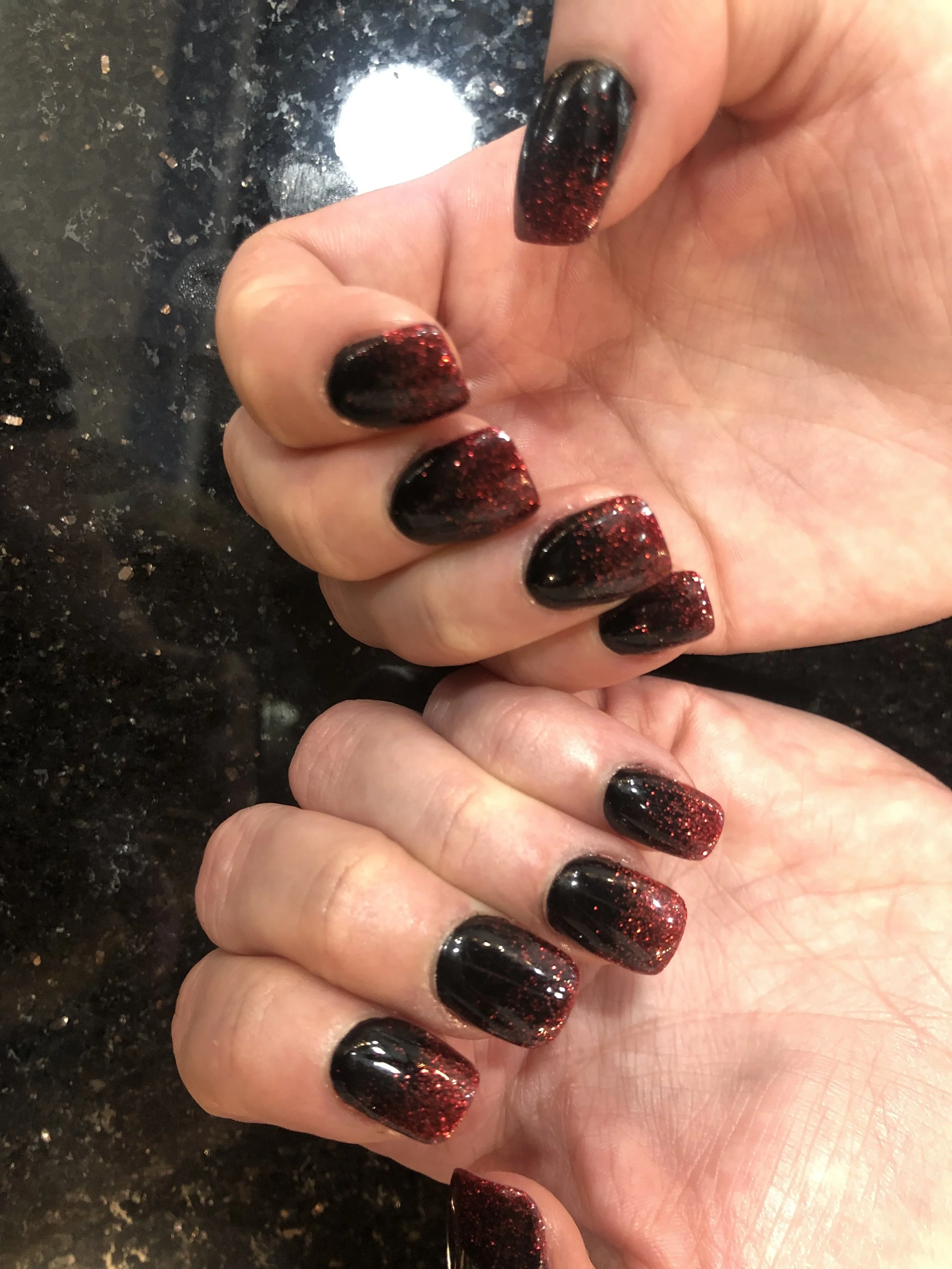 Close-up of a woman's hand displaying nails painted with black and red glitter polish, resting on a dark, textured surface.