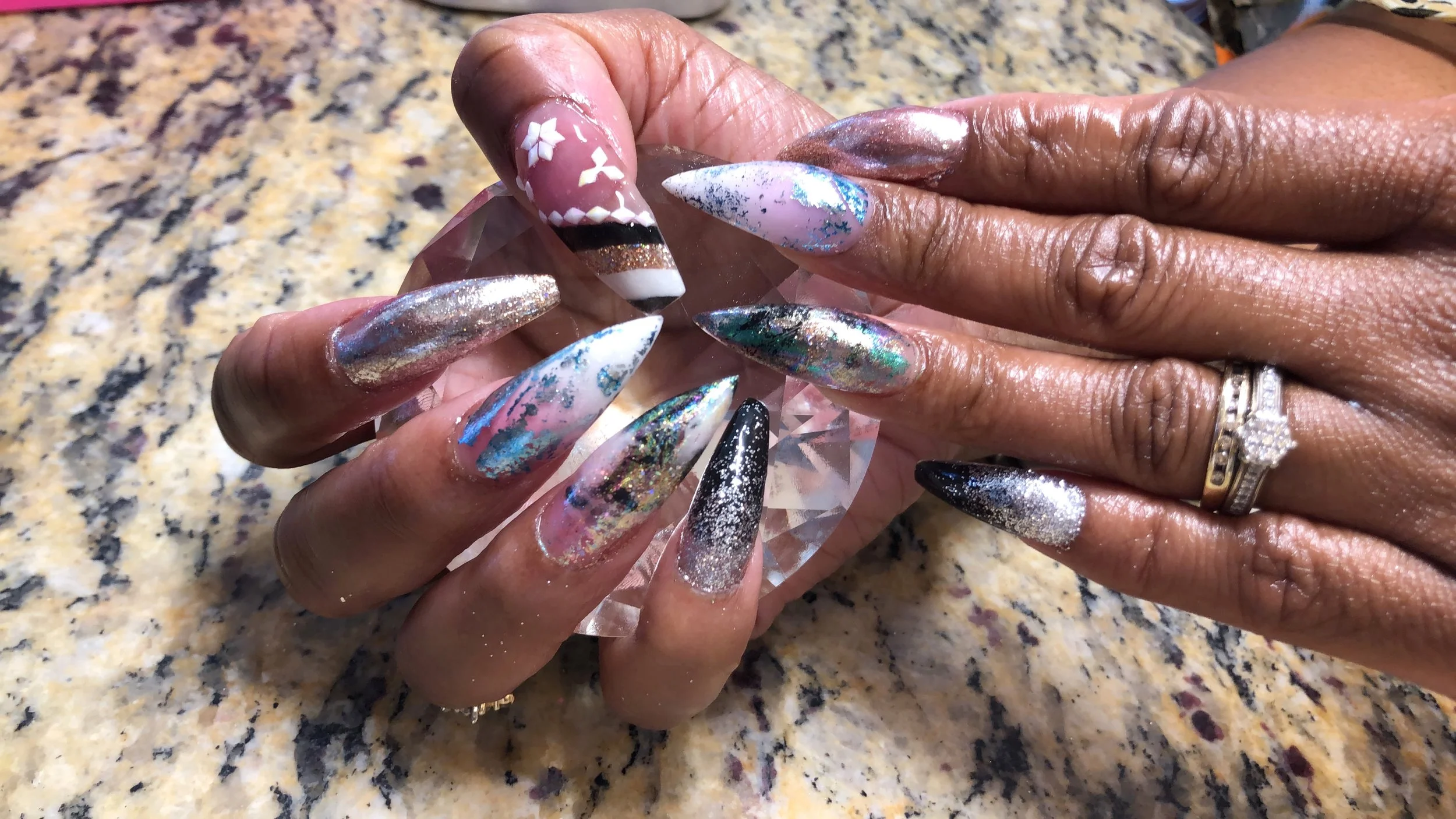 Close-up of hands with long, colorful, glittery, and holographic acrylic nails holding a faceted glass object on a speckled granite countertop.