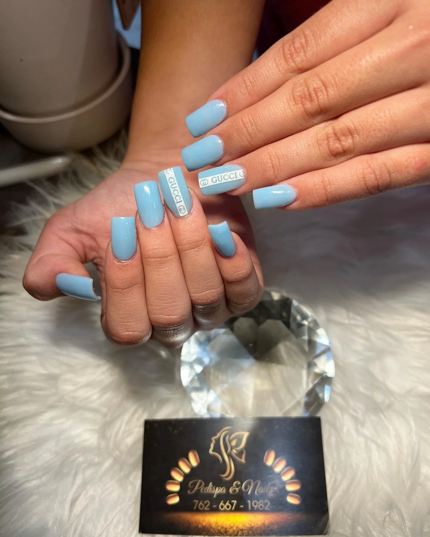 Close-up of light blue manicured nails with Gucci logo designs on two nails, holding a black and gold card with Cyrillic text and a decorative design, on a fluffy white surface with a reflective diamond-shaped ornament underneath.