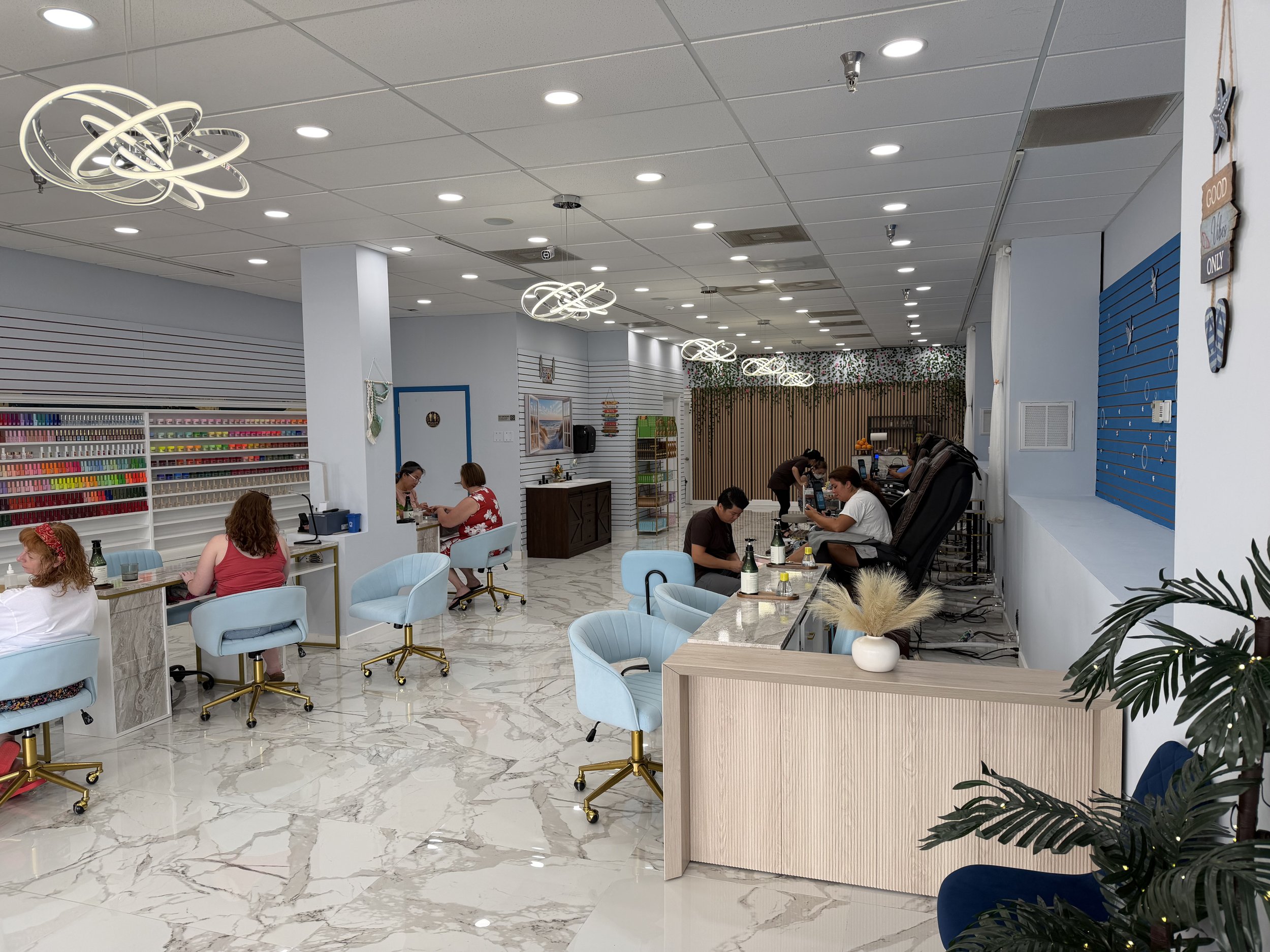 Interior of a nail salon with clients getting treatments, white and blue furniture, colorful nail polish display, modern lighting fixtures, and decorative plants.