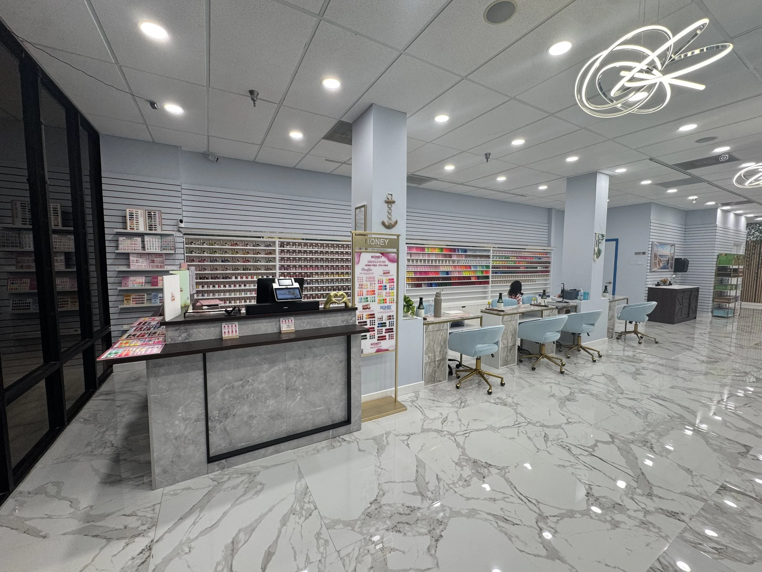 Interior of a retail store with glossy marble floors, white shelves stocked with colorful nail polish, a curved marble checkout counter, and seating area with light blue chairs, illuminated by circular ceiling lights and modern light fixtures.