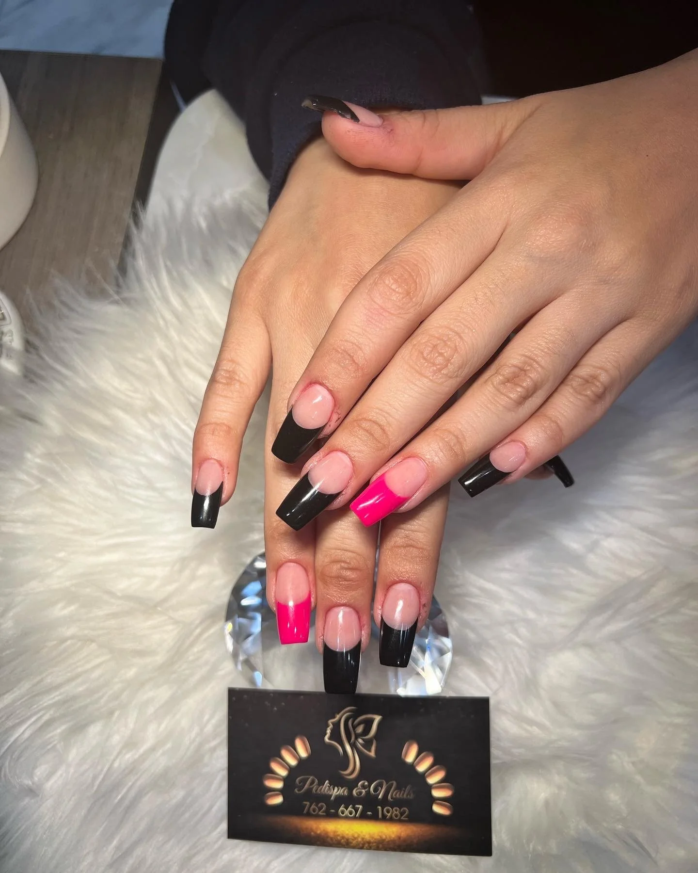 A woman's hands with freshly manicured nails, displaying black, pink, and natural nail designs, resting on a reflective surface with a business card for a nail salon nearby.
