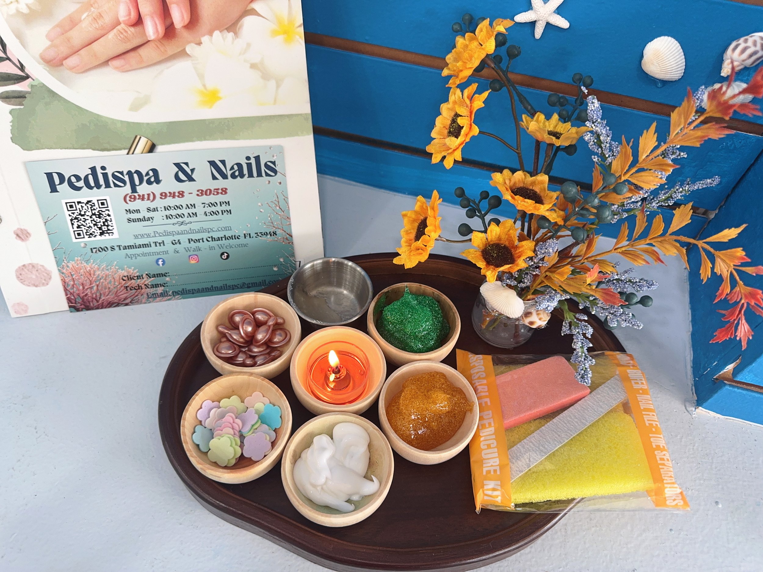 Decorative table display with artificial flowers, candles, and small containers filled with beads, slime, and foam shapes, set against a blue wall with seashells and seashell decoration, along with a sign for Pedispa & Nails.