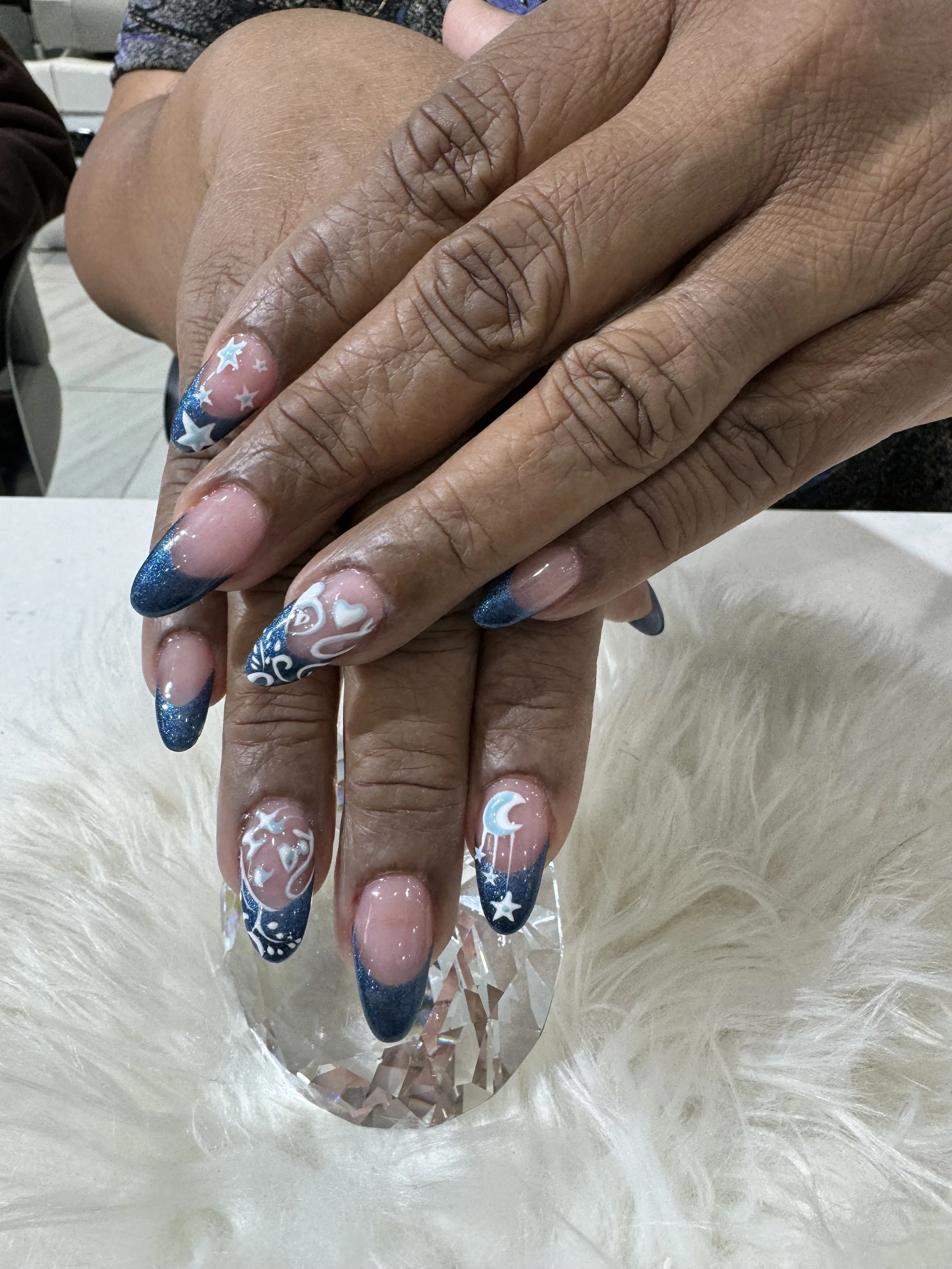Close-up of hands with long, decorated nails featuring a navy blue and white starry night design, resting on a faceted, transparent heart-shaped ornament, with a fuzzy white surface beneath and some background elements.