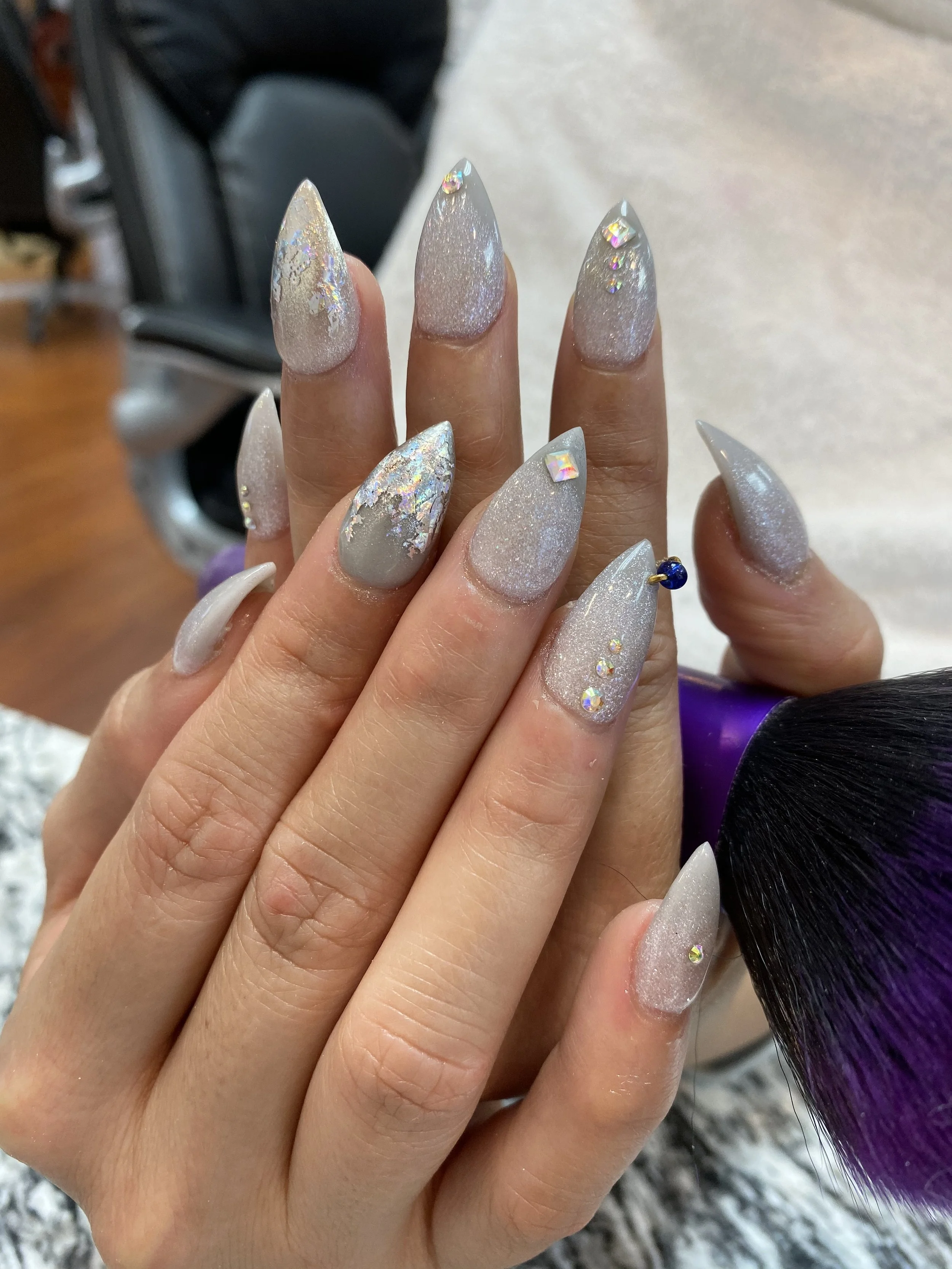 Close-up of a hand with long, almond-shaped, glittery, light-colored nails decorated with small rhinestones. In the background, blurred hair extensions and a purple hairbrush are visible.