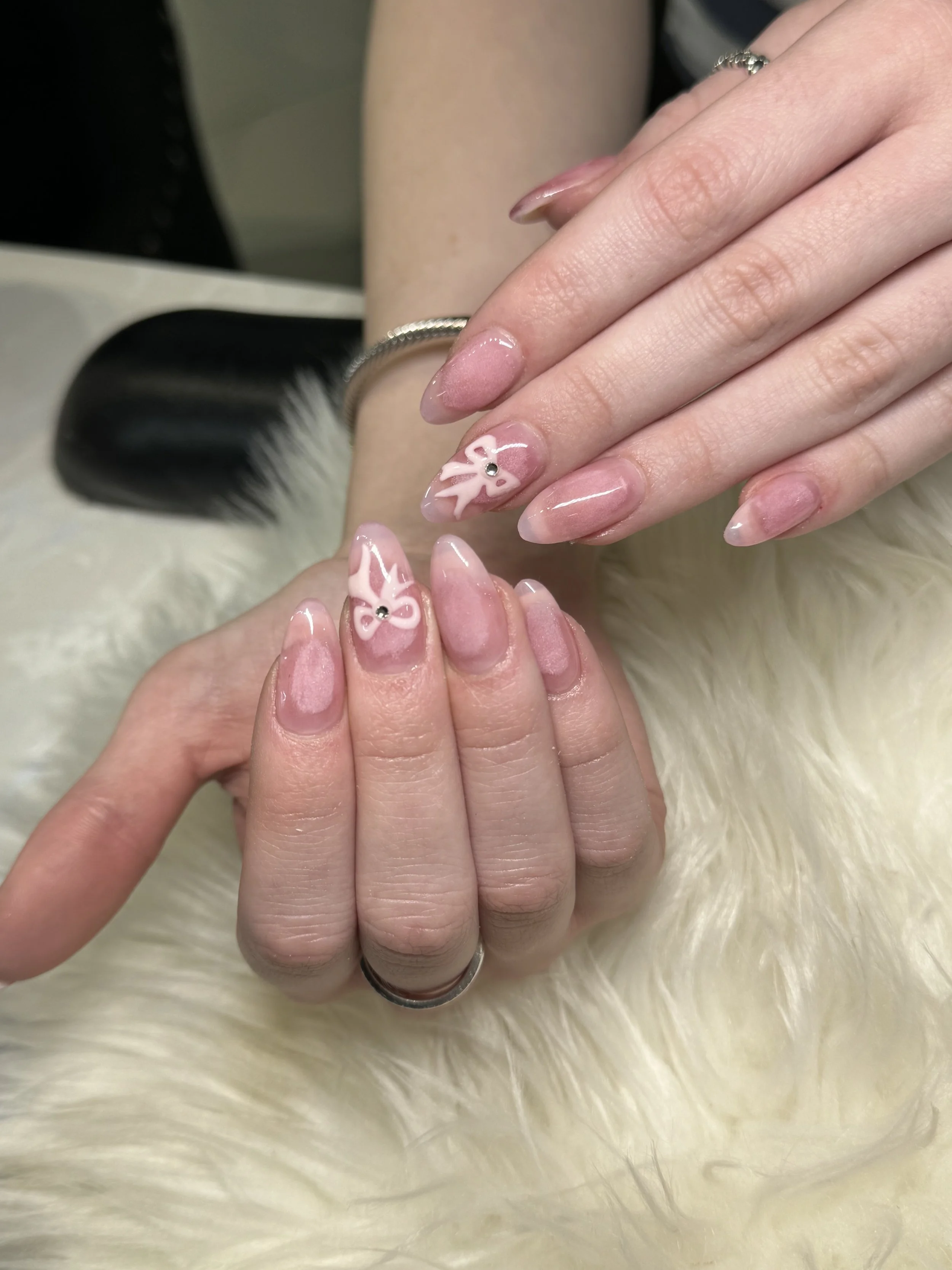 Close-up of a person's hand with manicured nails featuring pink and white floral designs with a small rhinestone in the center of each flower, resting on a soft, white, fluffy surface.