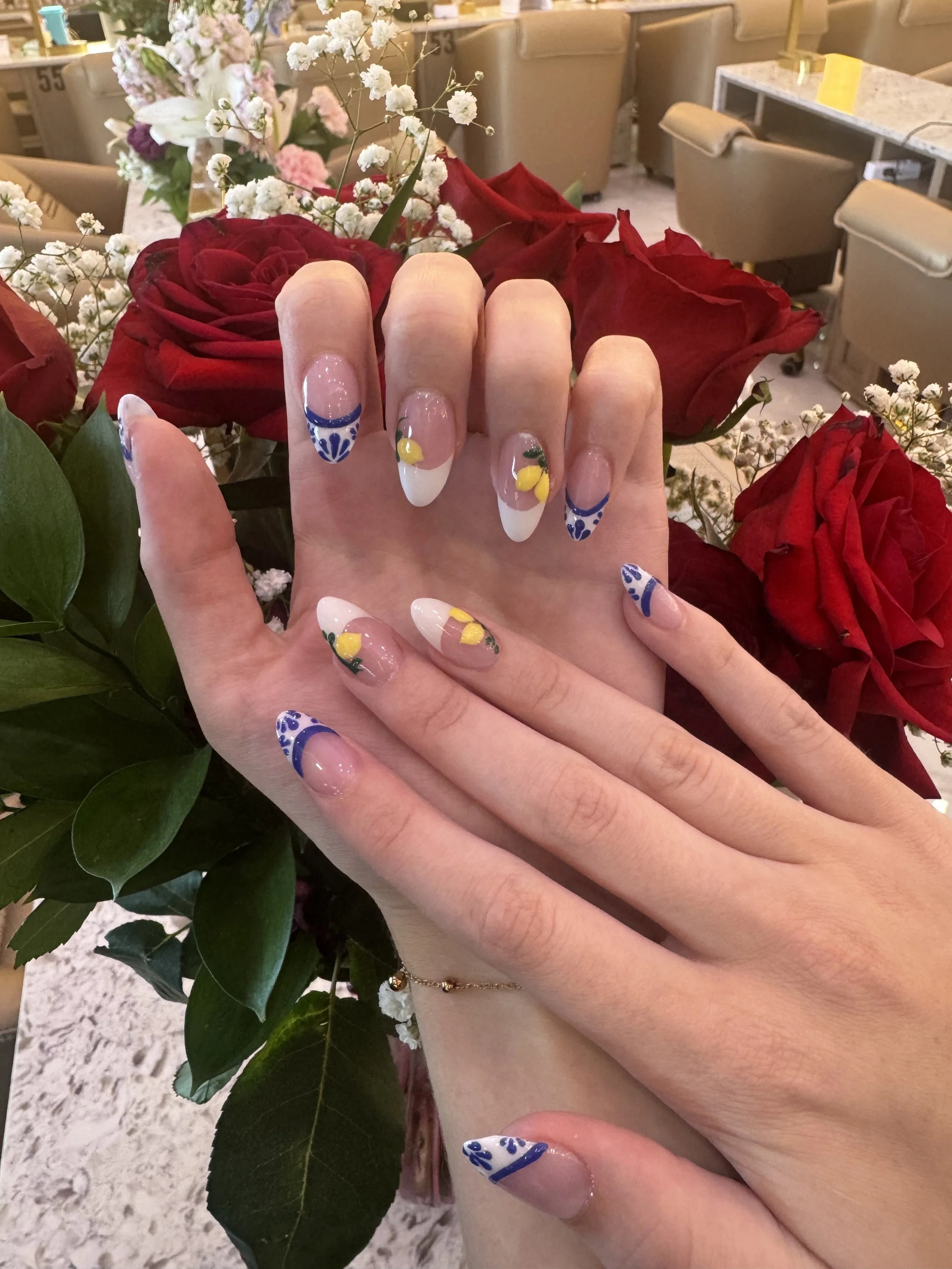 Close-up of a person's hand displaying decorated nails with a floral and lemon design, set against a bouquet of red roses and white baby's breath flowers.