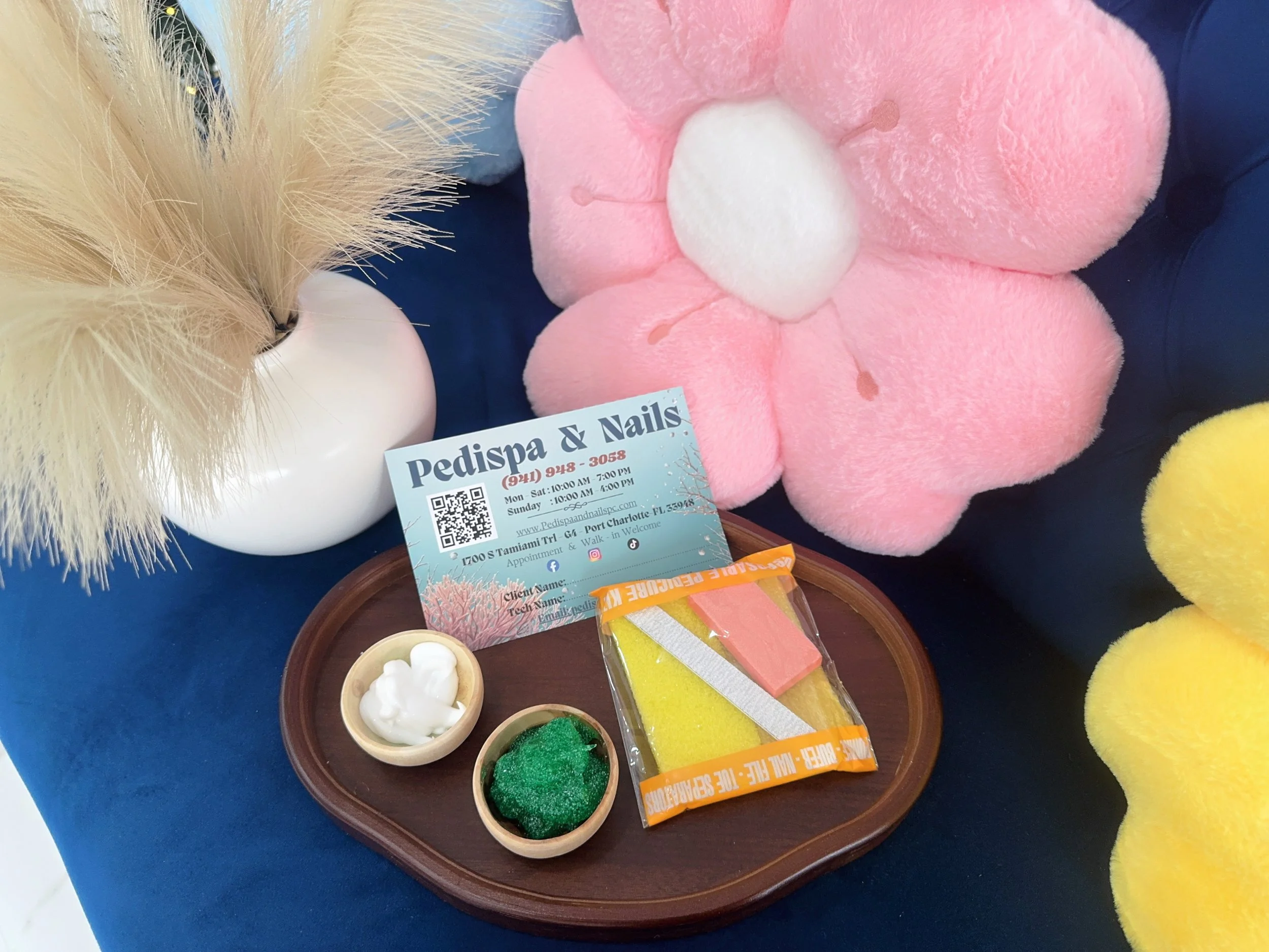 A display of pet grooming items including a pink plush flower-shaped item, a white plush ball, a pink and white apple-shaped toy, small bowls with white and green grooming products, a business card for Pedispa & Nails, and colorful professional spong