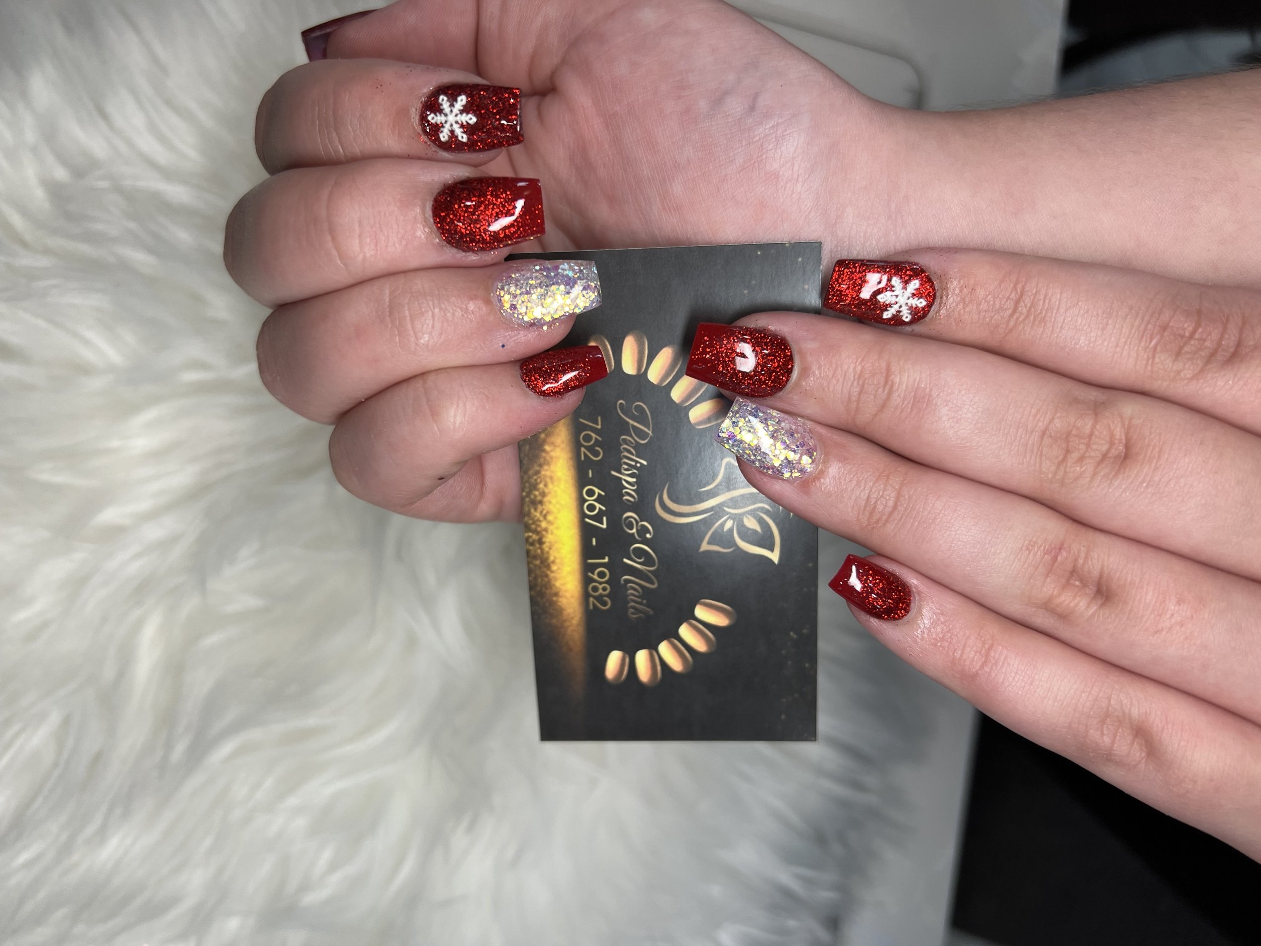 Close-up of a person's hand showing red glitter nail polish with white snowflake design on some nails and holographic glitter nail polish on others, holding a black business card.