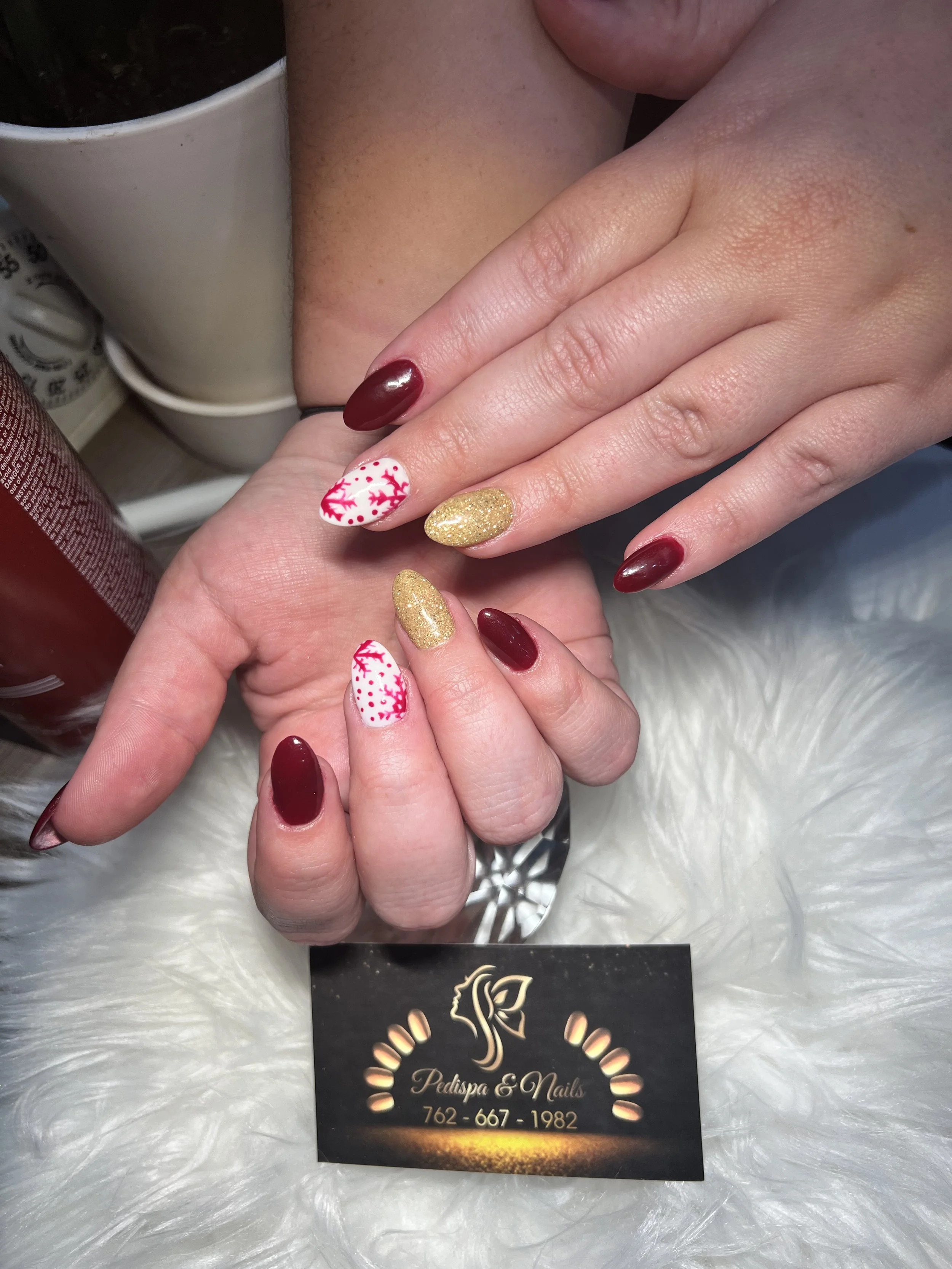 Close-up of a person's manicured hand resting on another hand, with decorated nails featuring red, white, and gold colors, in front of a business card for Pedispa & Nails, on a white furry surface.