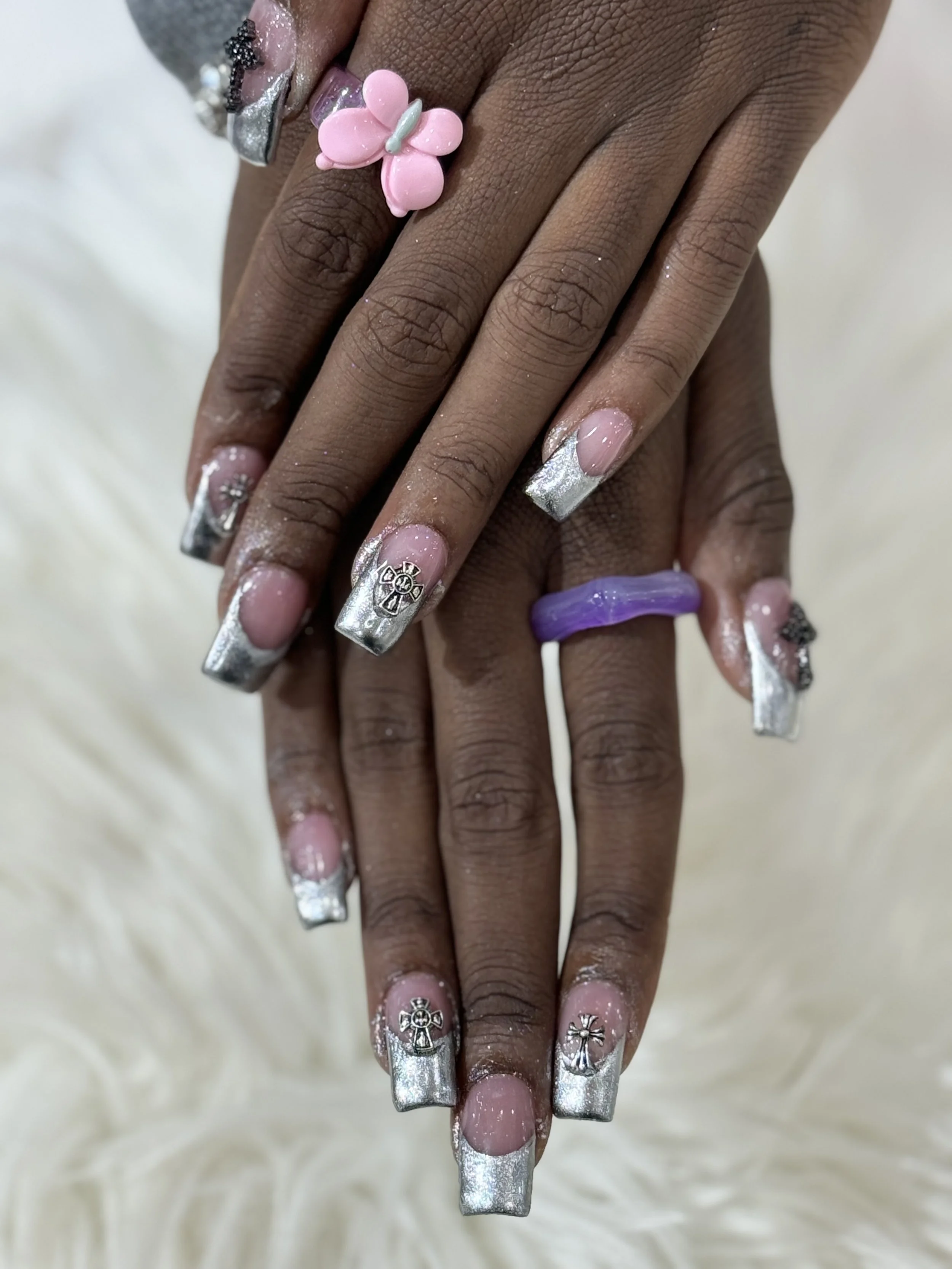 Close-up of a person's hand with long, metallic silver nails decorated with small angel cross and other religious charms, and a pink butterfly nail on the finger, against a soft, blurred background.