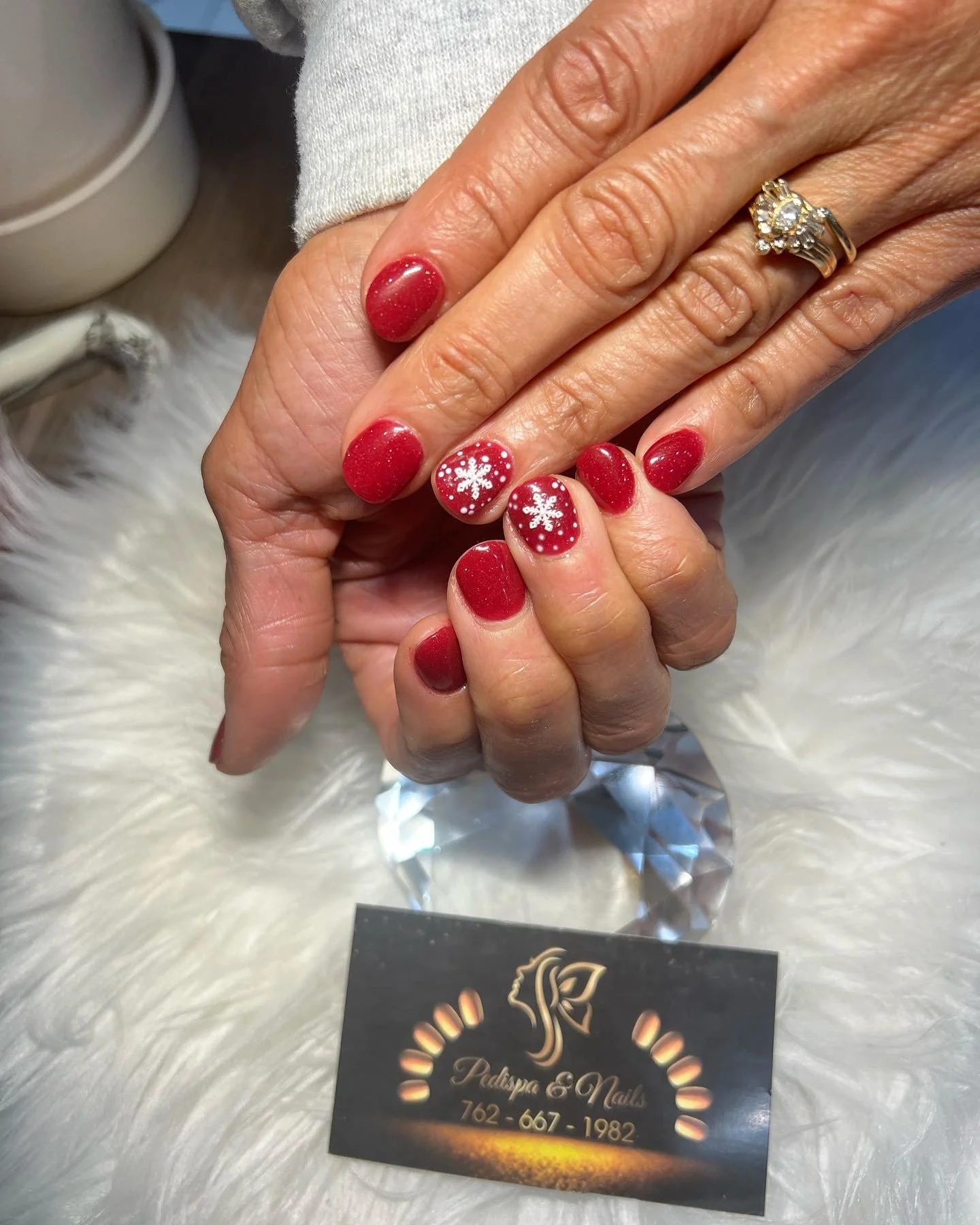 Close-up of a woman's hand with red Christmas-themed nail art and a sparkling ring, resting on a business card on a fluffy white surface.