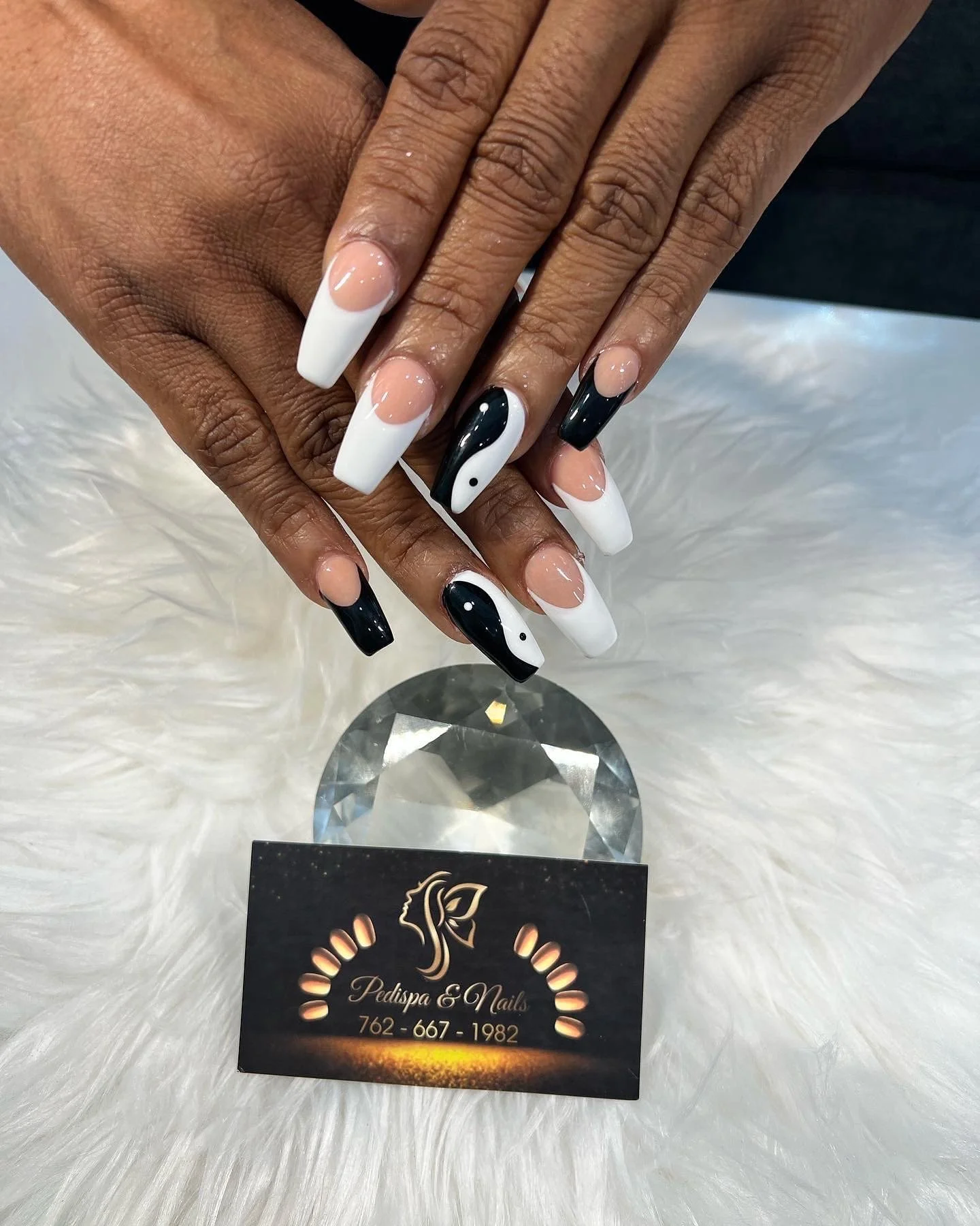 Hands with manicured nails resting on a surface, showing black and white nail designs. A business card for Pedispa & Nails is placed below, with a phone number. The background is white fur-like material.